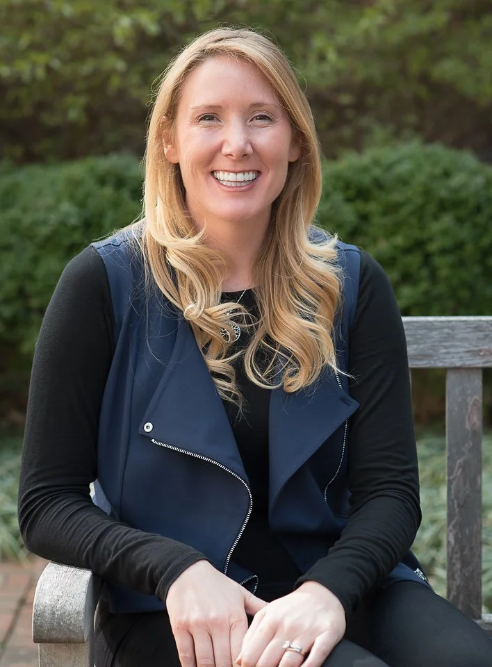 Young woman with long blonde hair smiling, sitting on a wooden bench outdoors, wearing a black top and a navy vest.