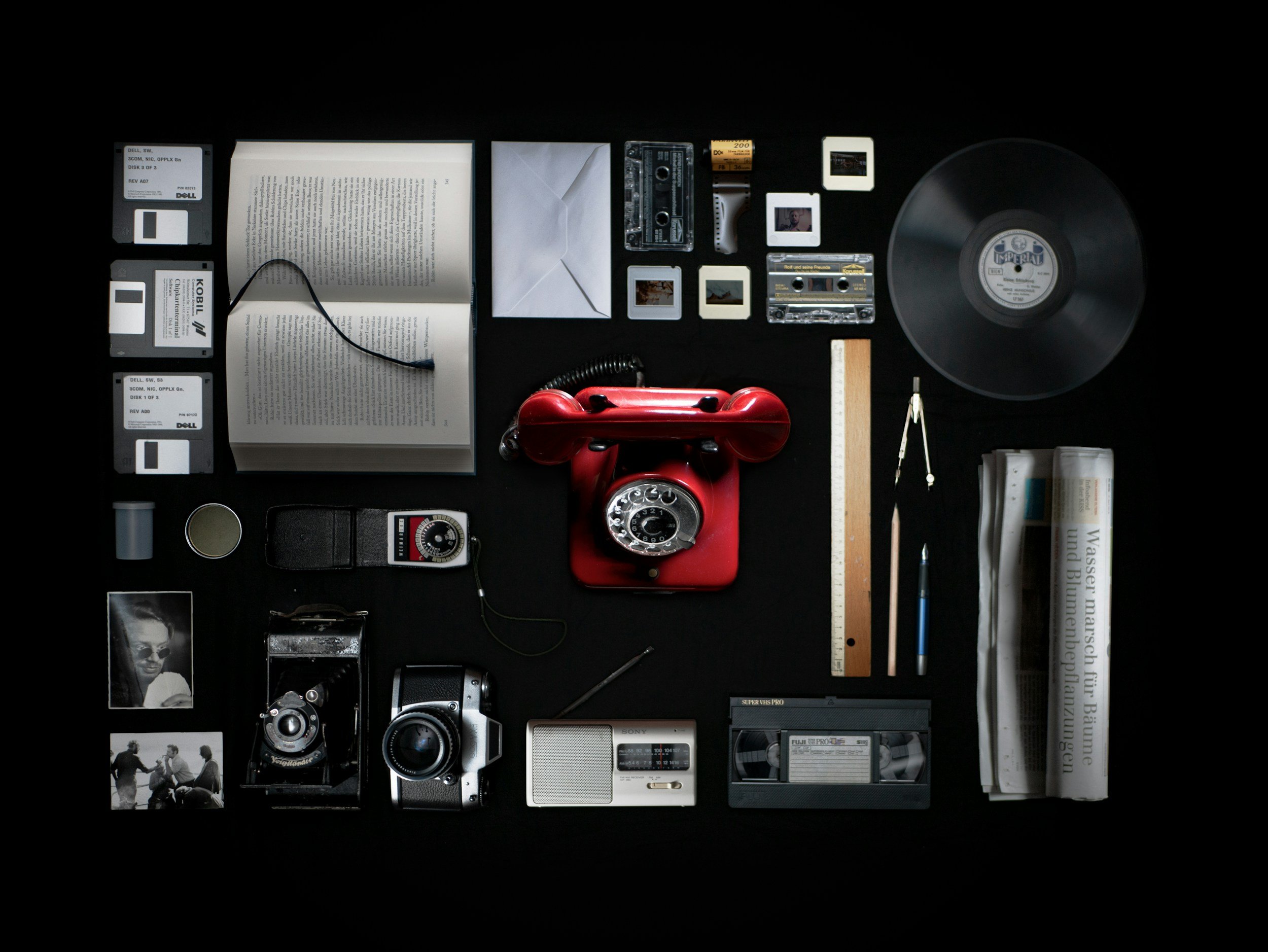 A flat lay of various vintage objects on a black background, including floppy disks, a book, a red rotary phone, photographs, a vinyl record, camera equipment, a newspaper, and writing supplies.