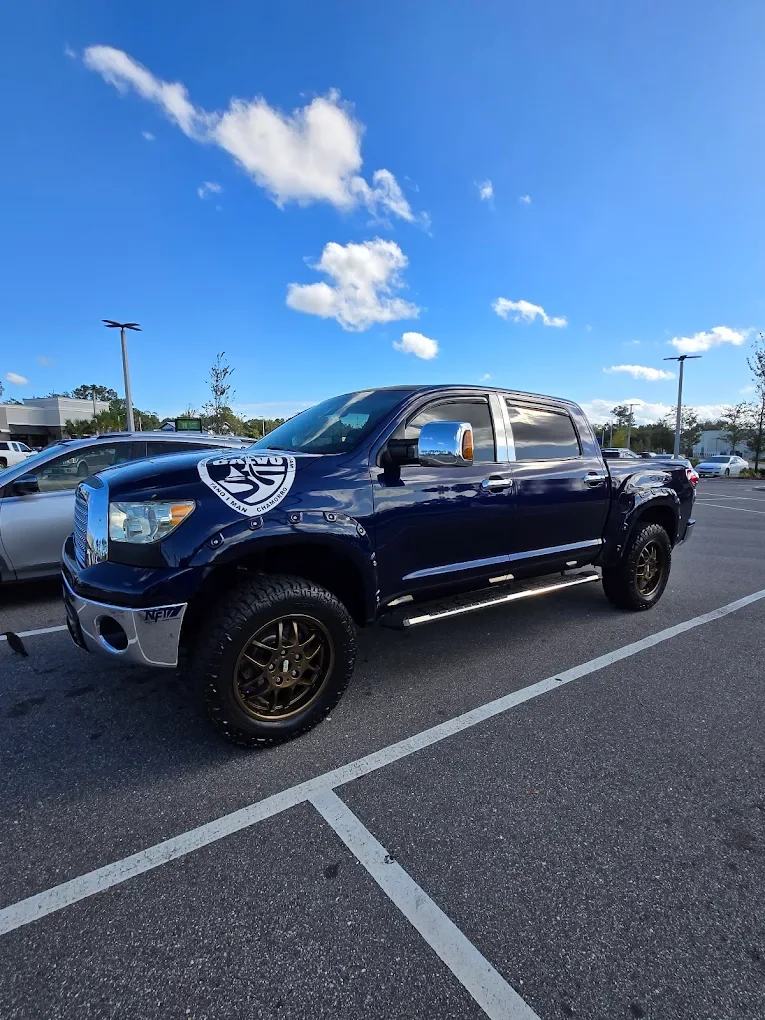 A black pickup truck with large wheels parked in a shopping plaza parking lot under a blue sky with scattered white clouds after being detailed by Coffee N Cars.