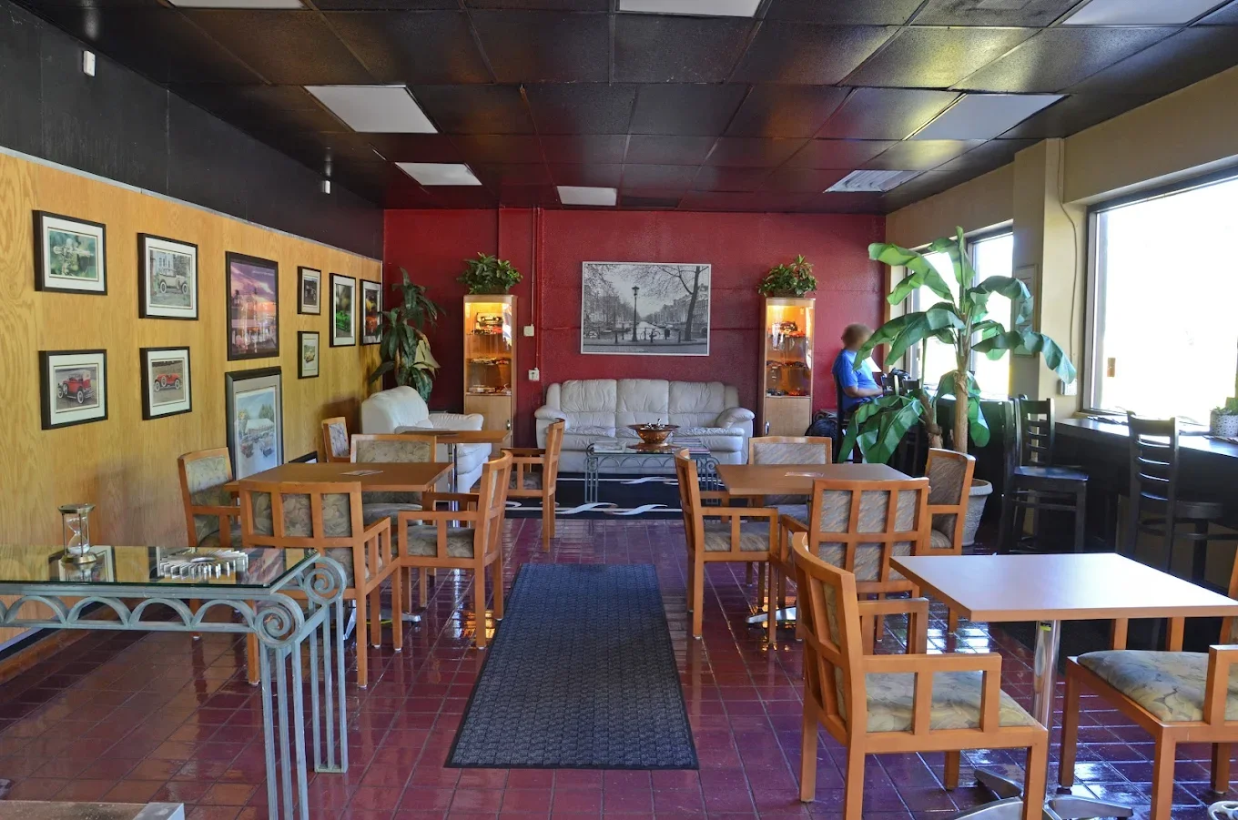 Inside of Coffee N Cars is  a cozy cafe with wooden tables and chairs, framed pictures on the wall, a sofa against a red accent wall, and a person standing near the window.