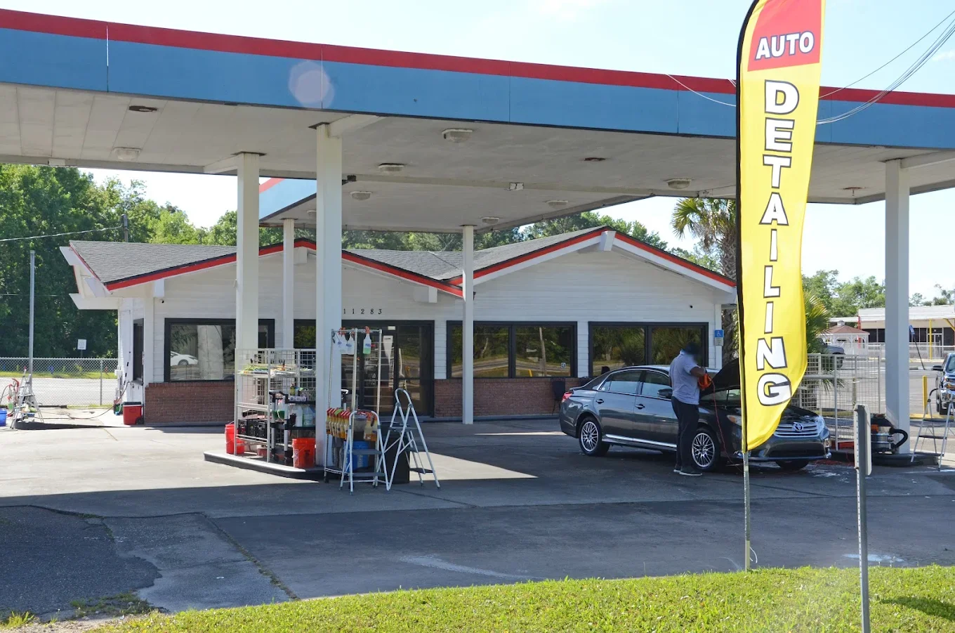 A gas station with a food or retail store, parked cars, and a bright yellow flag sign that says 'Auto Detailing.' A person is standing next to a car with the hood open, possibly working on it.
