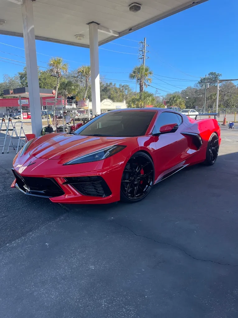 Red sports car parked under a canopy at a gas station with palm trees and utility poles in the background.
