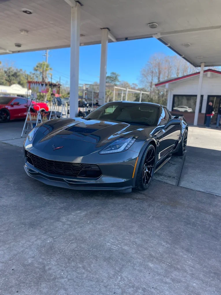 A dark gray Chevrolet Corvette sports car parked outside a building with a covered area, with another red sports car visible in the background.