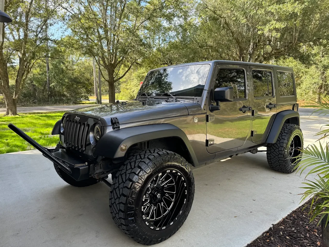 Black Jeep Wrangler with large tires parked on concrete driveway, surrounded by trees and grass in a sunny outdoor setting. Detailed by Coffee N Cars.