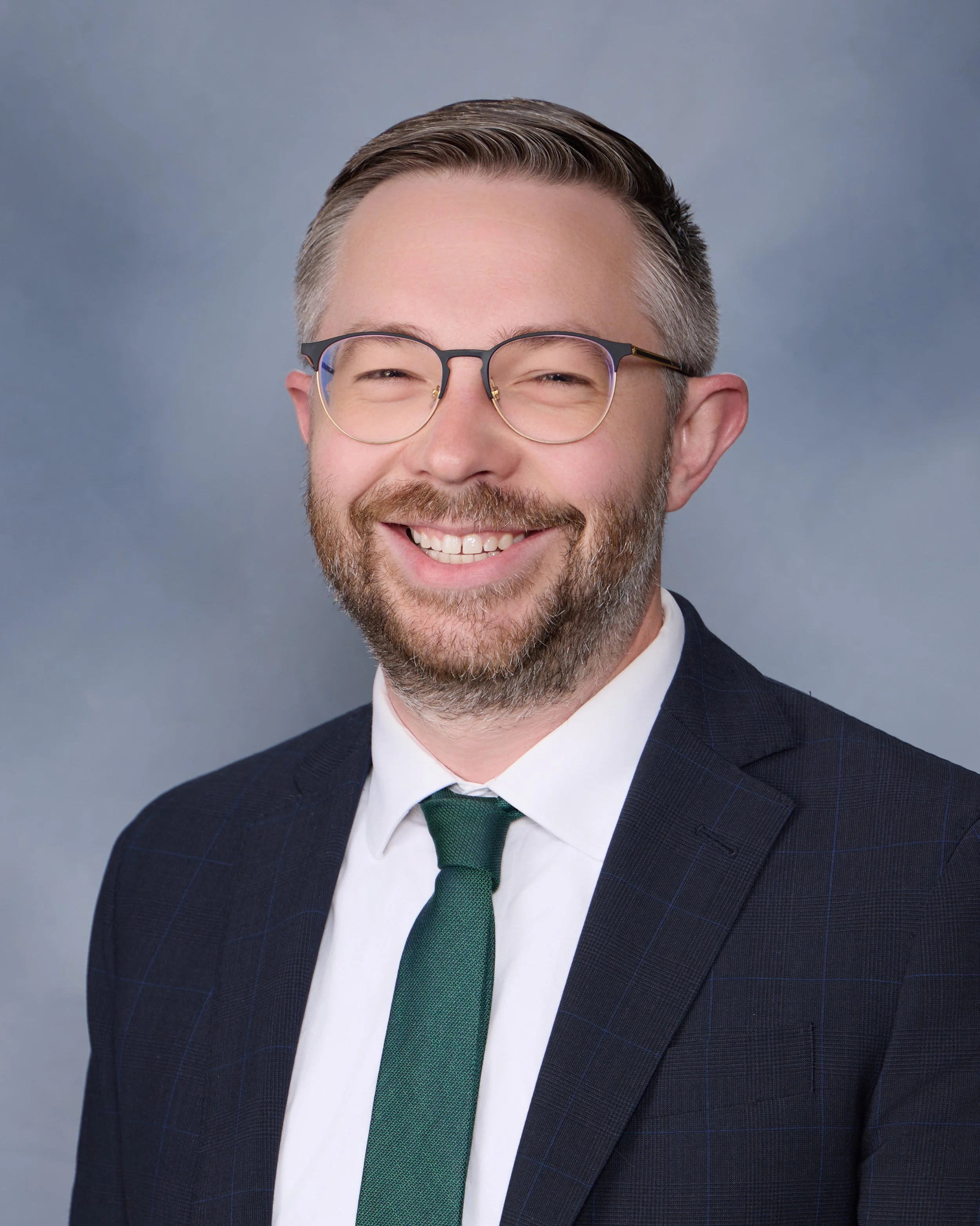 Professional headshot of a smiling man with glasses, a beard, wearing a suit jacket, white shirt, and green tie, against a gray background.