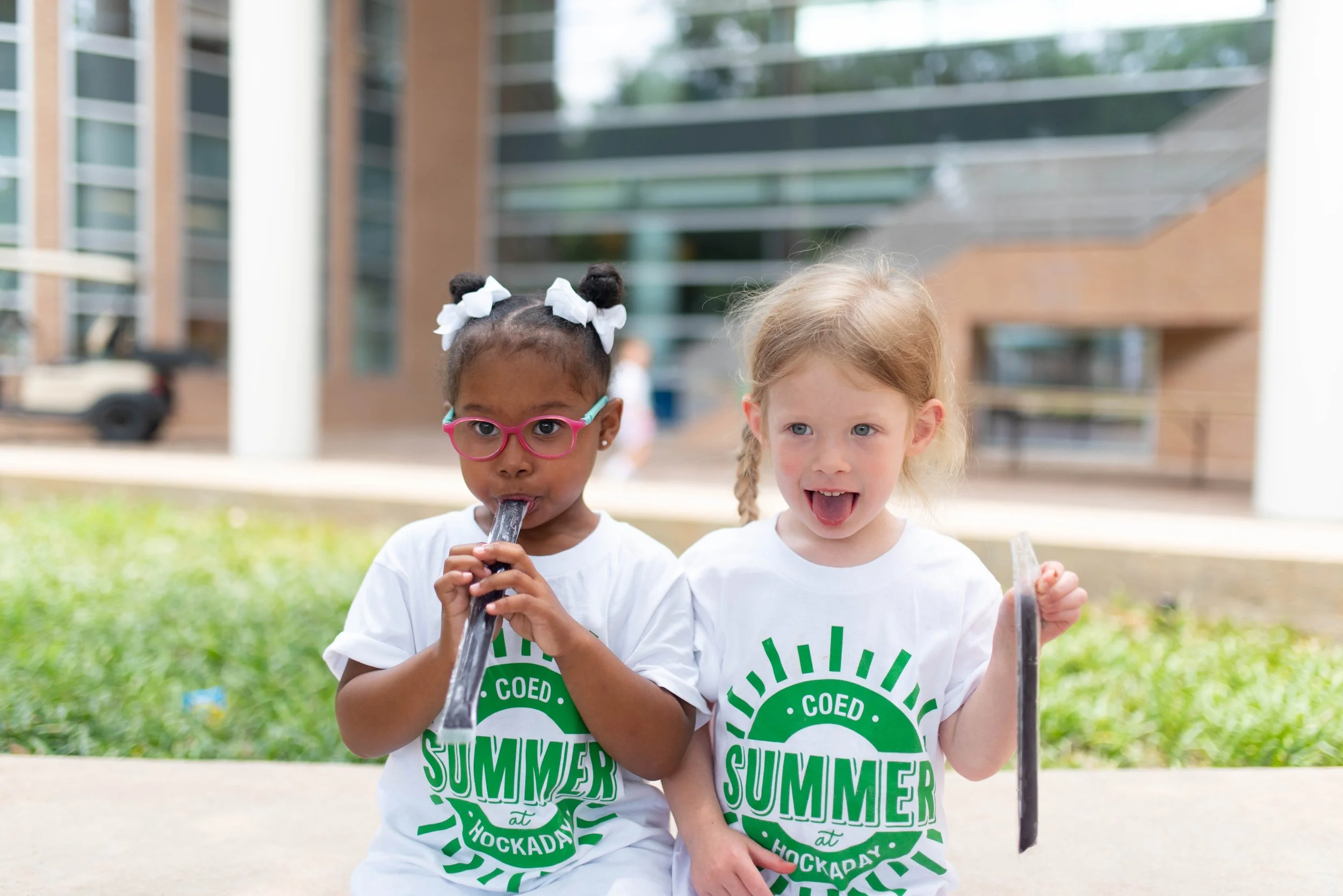 Two young girls sitting outdoors, wearing white T-shirts with green text, enjoying ice pops. The girl on the left has dark hair in buns with white bows, glasses, and is licking her ice pop, while the girl on the right has light hair in a braid, sticking out her tongue and holding her ice pop. Background shows a modern building and greenery.