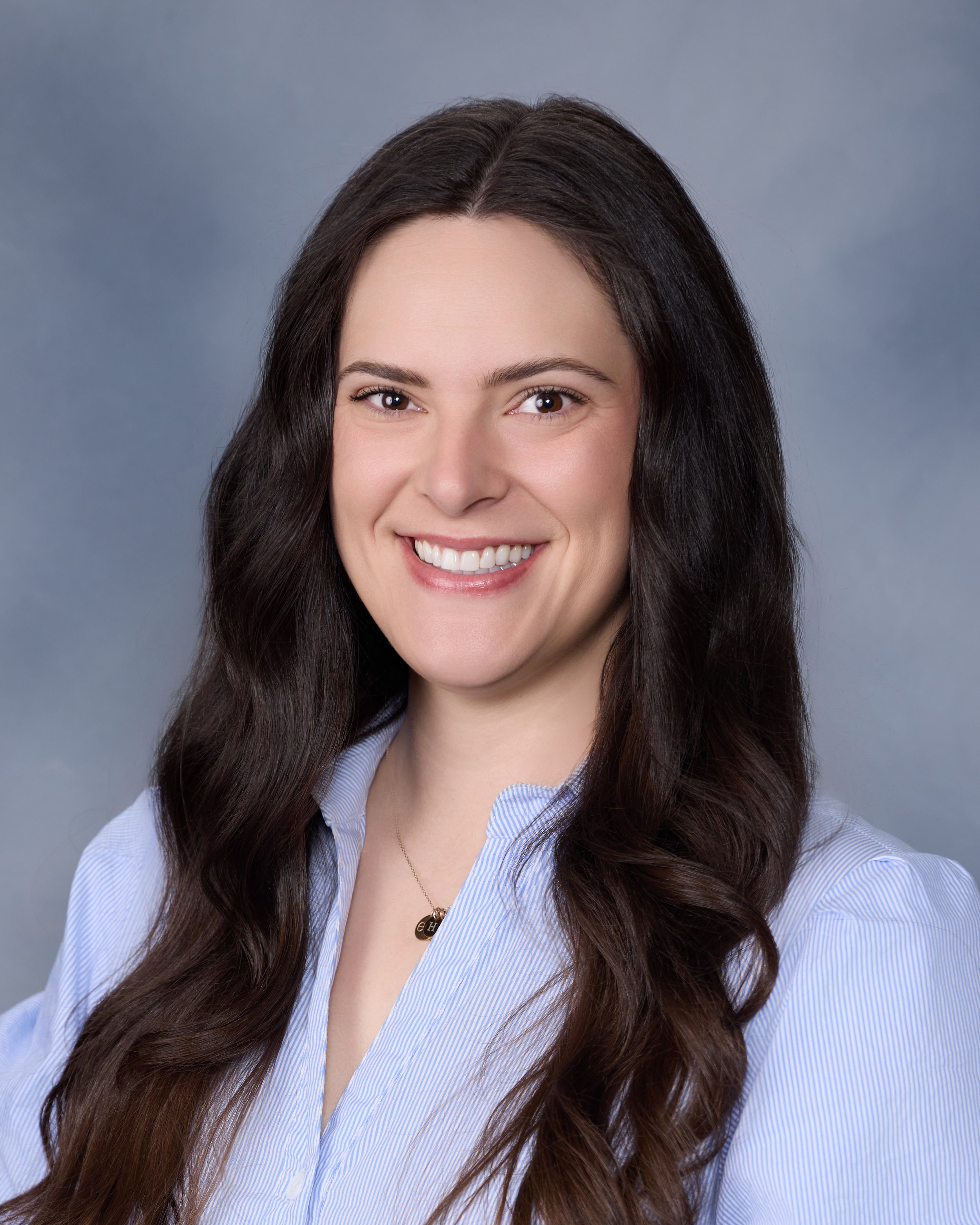 A woman with long dark brown hair, smiling, wearing a light blue shirt and a gold necklace, against a gray background.