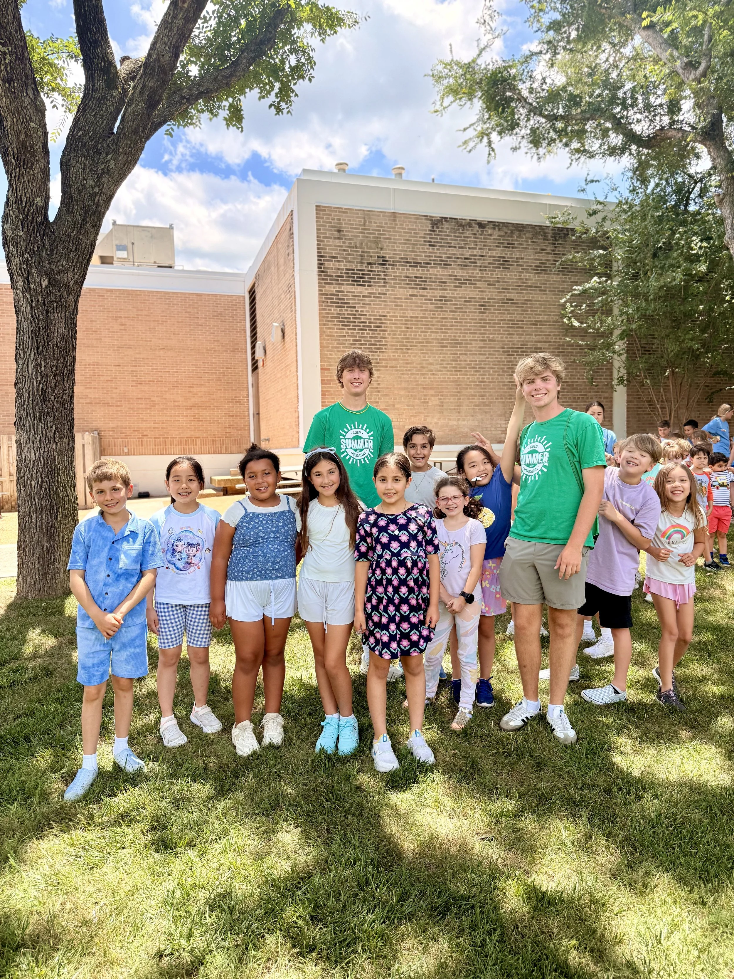 Group of children and two young men standing outdoors on a sunny day, smiling, in front of brick building and trees.
