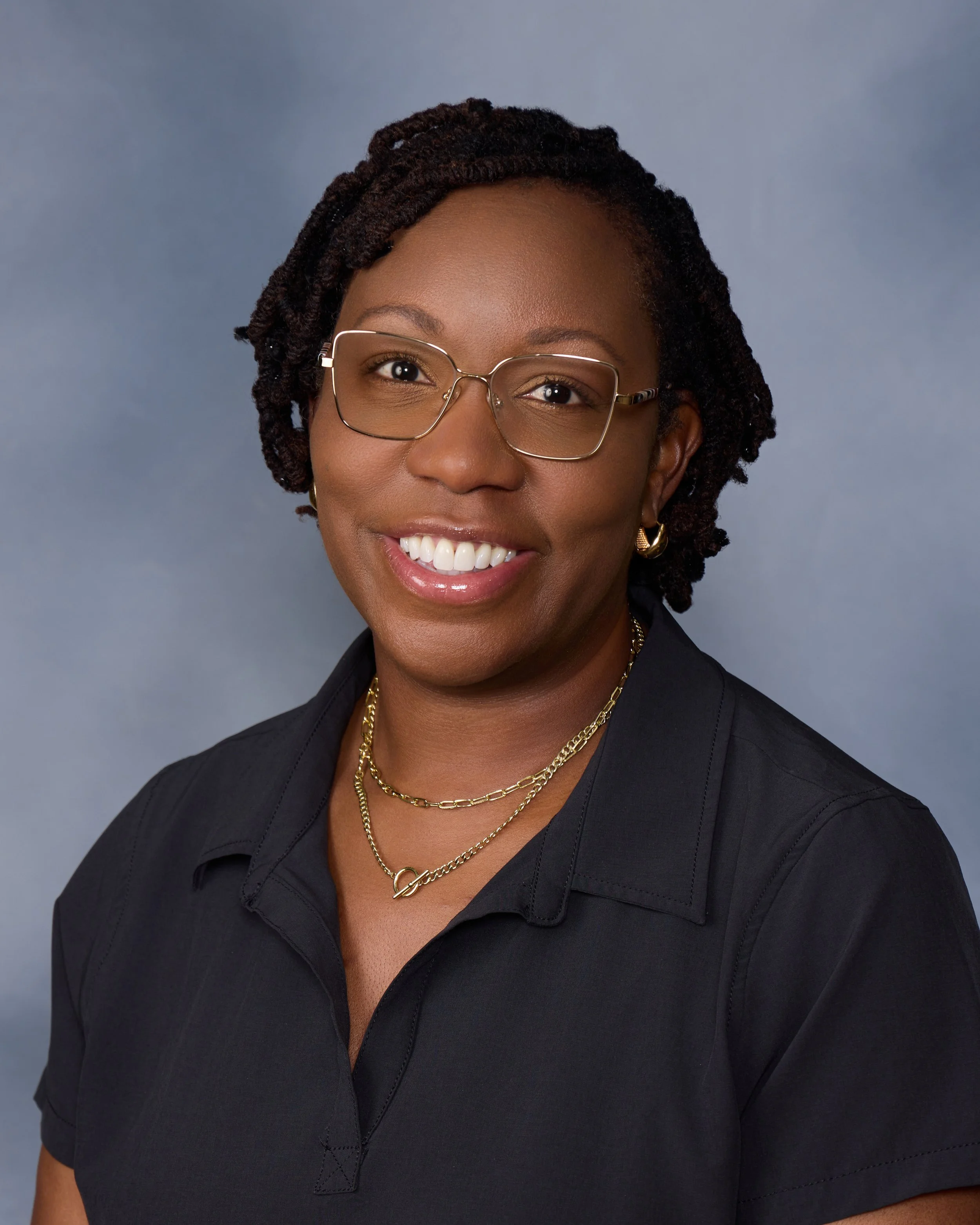 A portrait of an African American woman wearing glasses, gold jewelry, and a black shirt, smiling against a gray background.