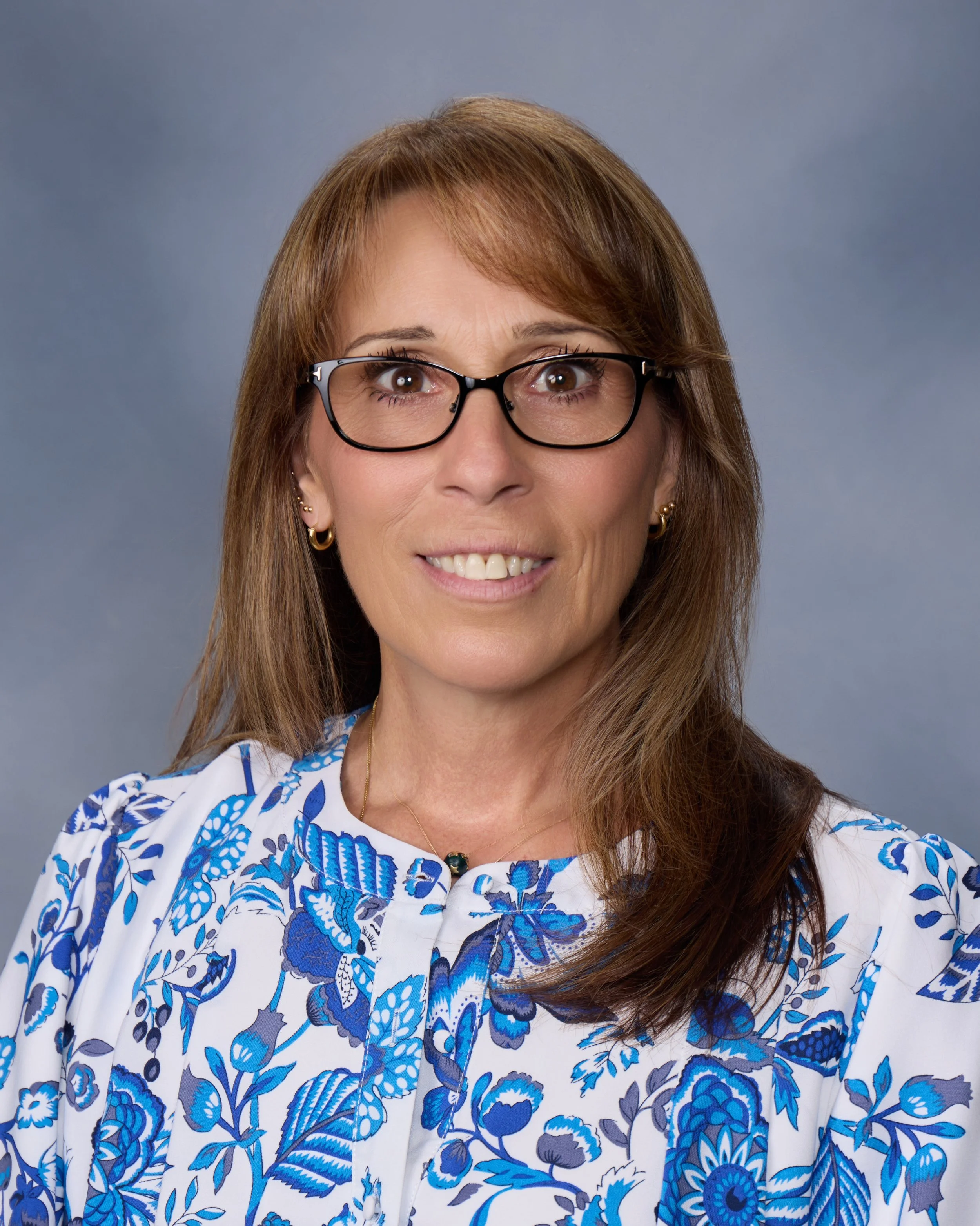 Portrait of a woman with brown hair, glasses, wearing a white blouse with blue floral pattern, earrings, and a necklace against a gray background.