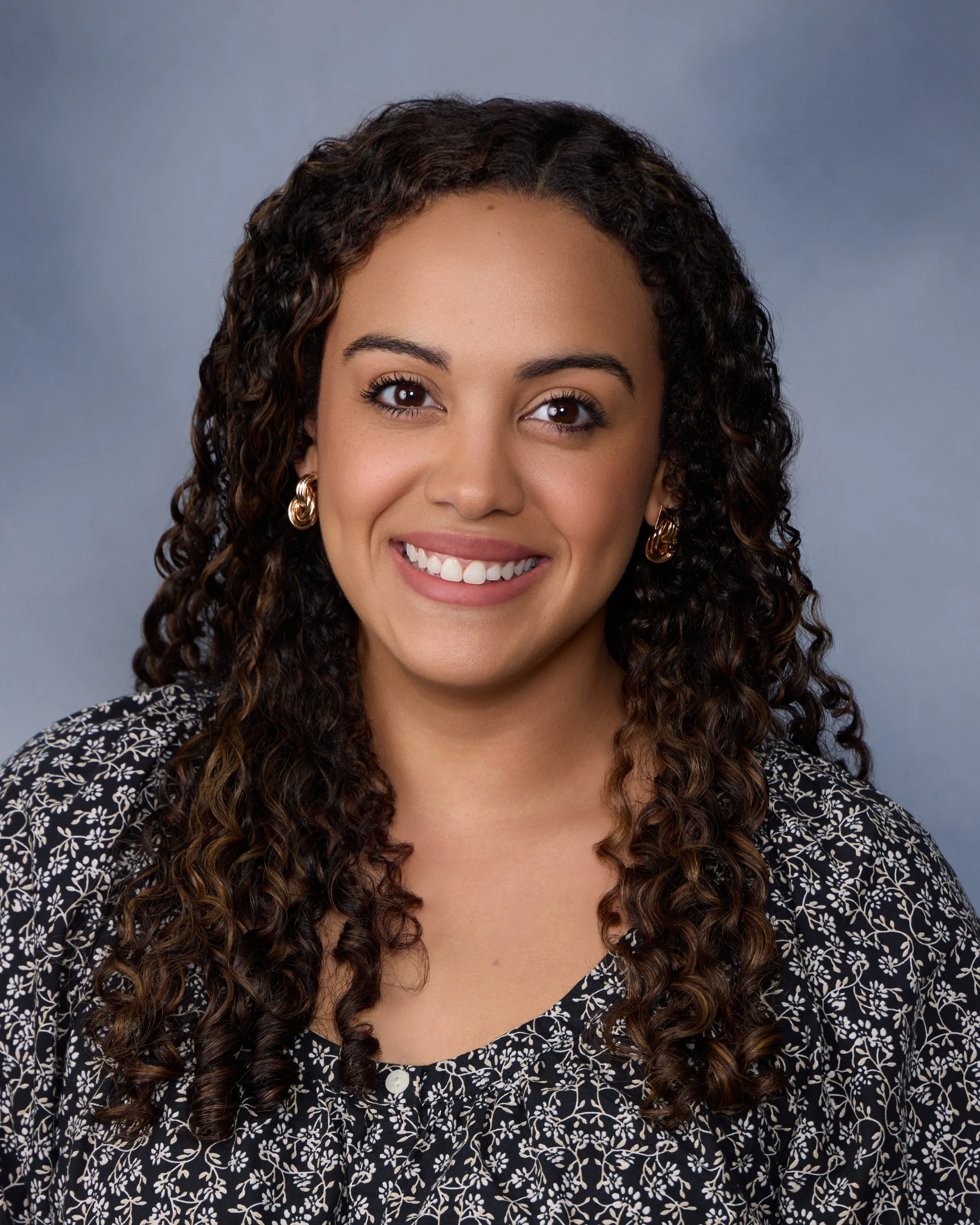 Close-up portrait of a woman with dark curly hair, wearing gold hoop earrings and a patterned blouse, smiling against a gray background.