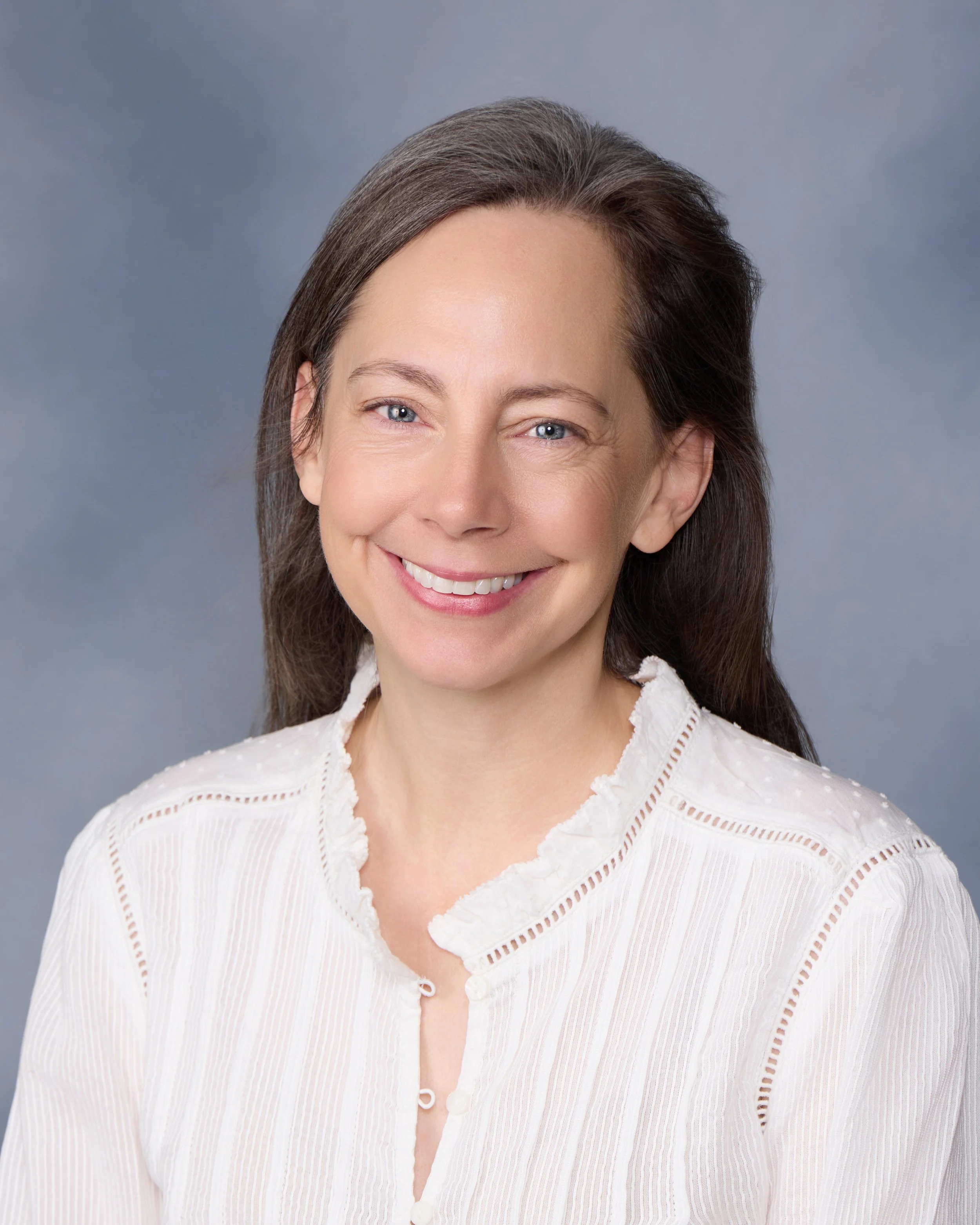 Headshot of a smiling woman with blue eyes and long brown hair, wearing a white blouse, against a gray background.