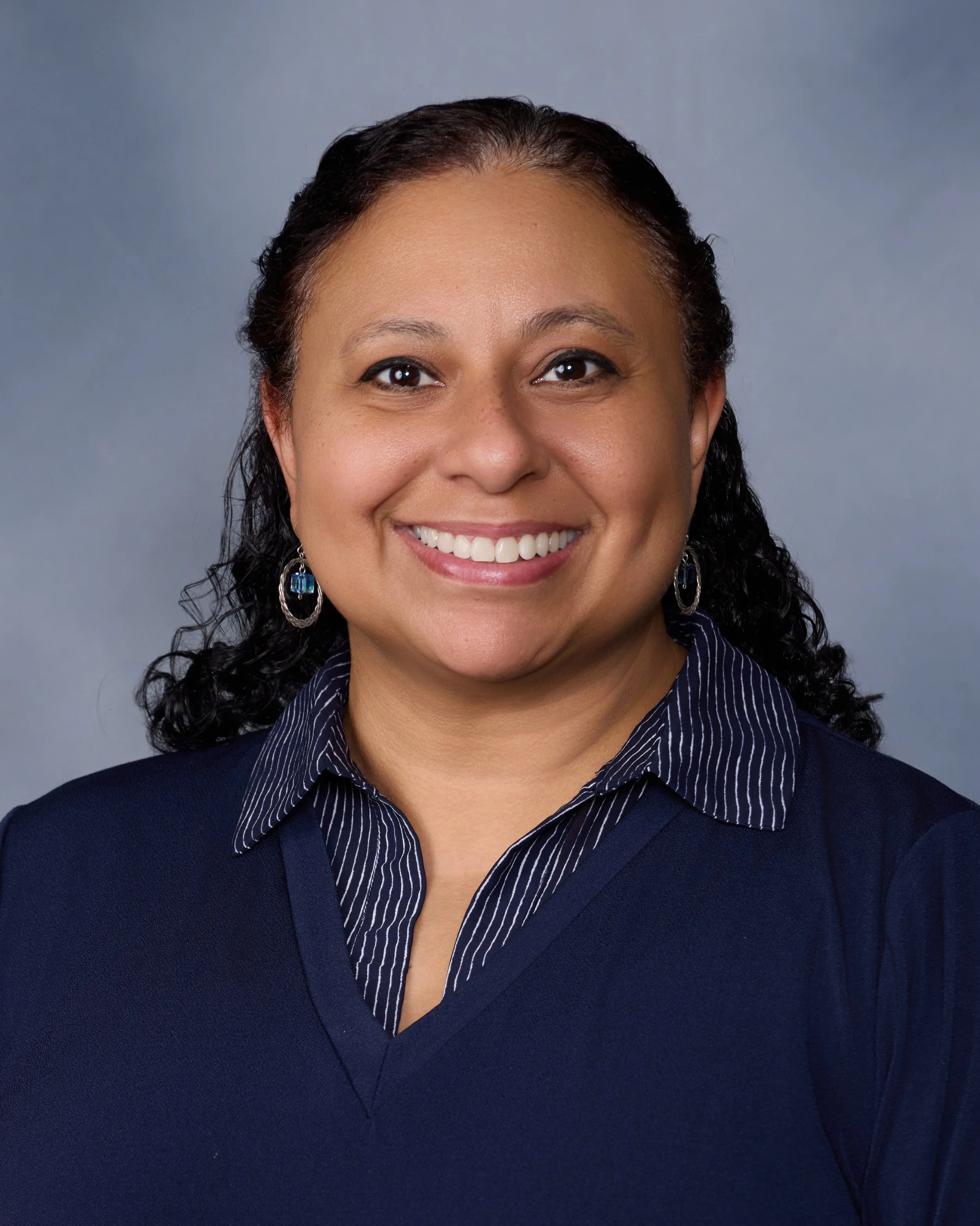 A woman with dark curly hair, smiling, wearing a dark blue sweater and earrings, against a gray background.