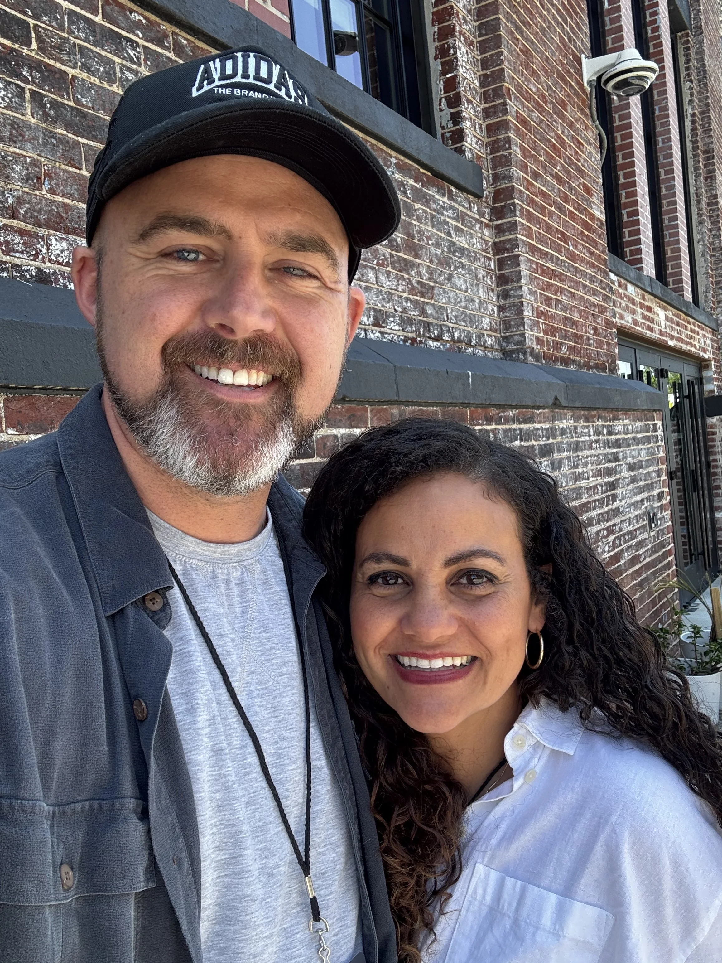 A smiling man and woman taking a selfie outside in front of a brick building.