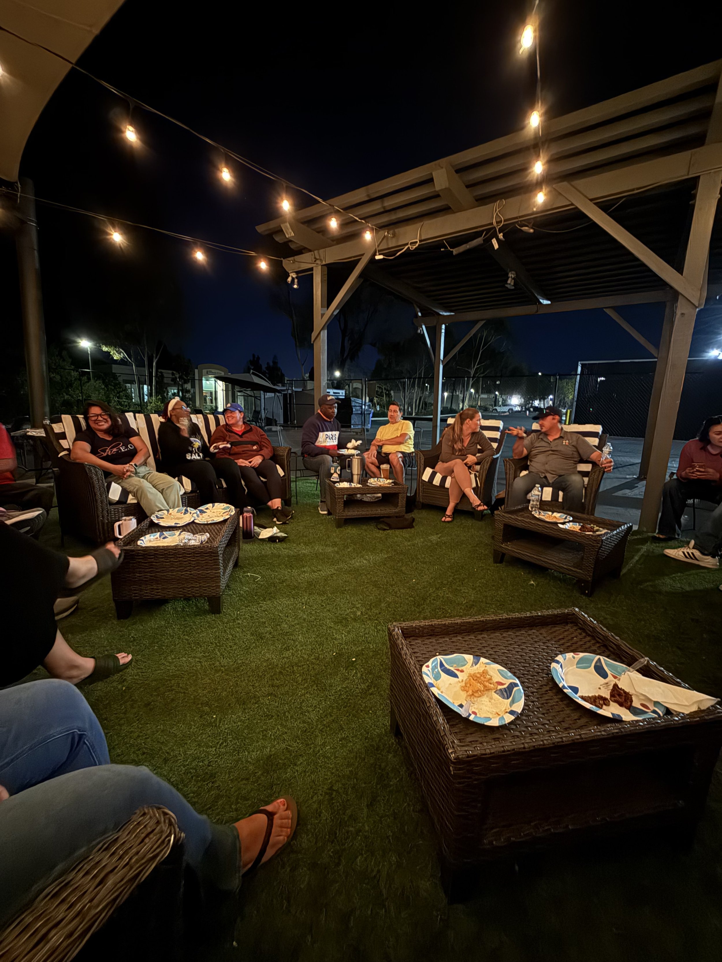 People sitting around a bonfire pit at night, socializing under string lights in an outdoor area with a covered patio.