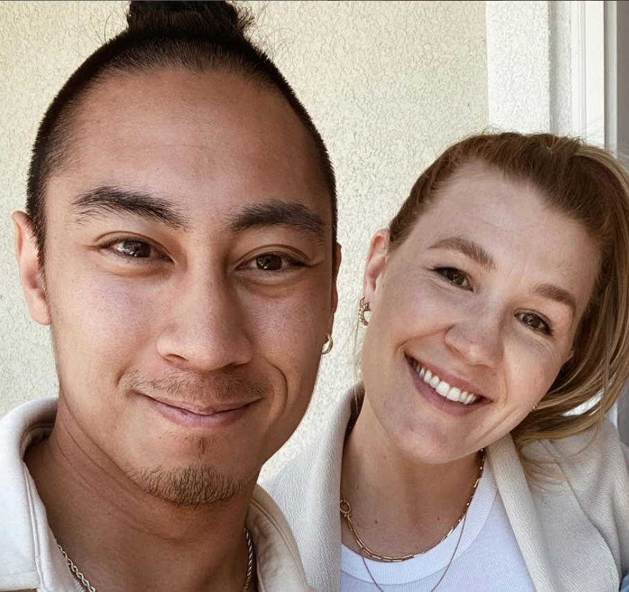 Matt and Esmae Villa are the Junior High, 6th-8th grade pastors at Carmel Church in San Diego. Close-up of smiling young man and woman taking a selfie outdoors near a building wall, both wearing light-colored clothing and gold jewelry.