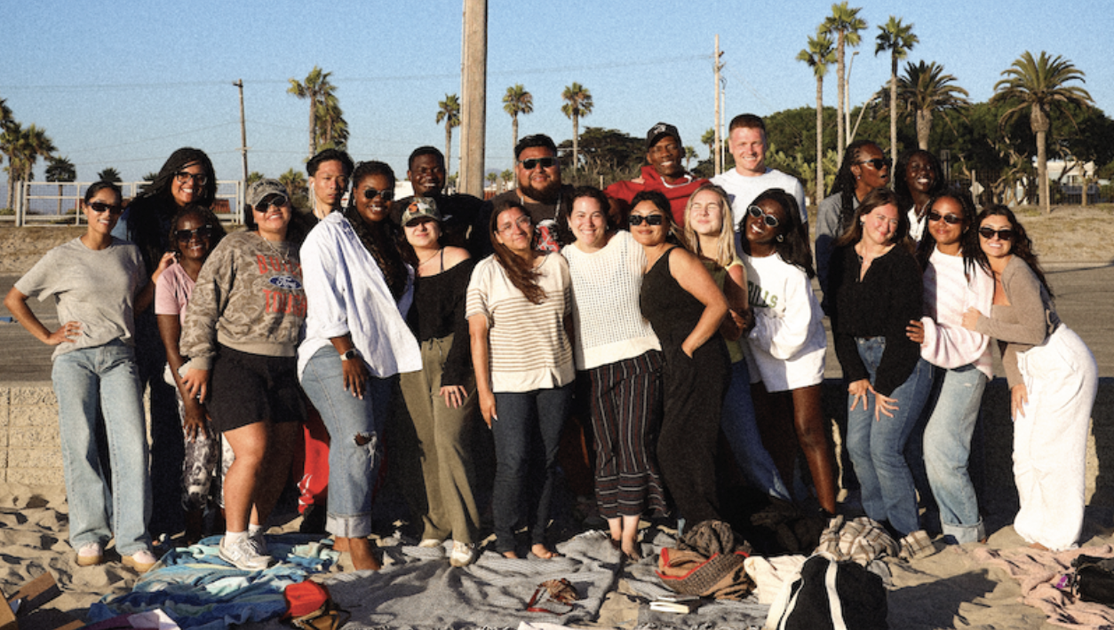 Group of diverse people smiling and posing for a photo on a beach with palm trees in the background.