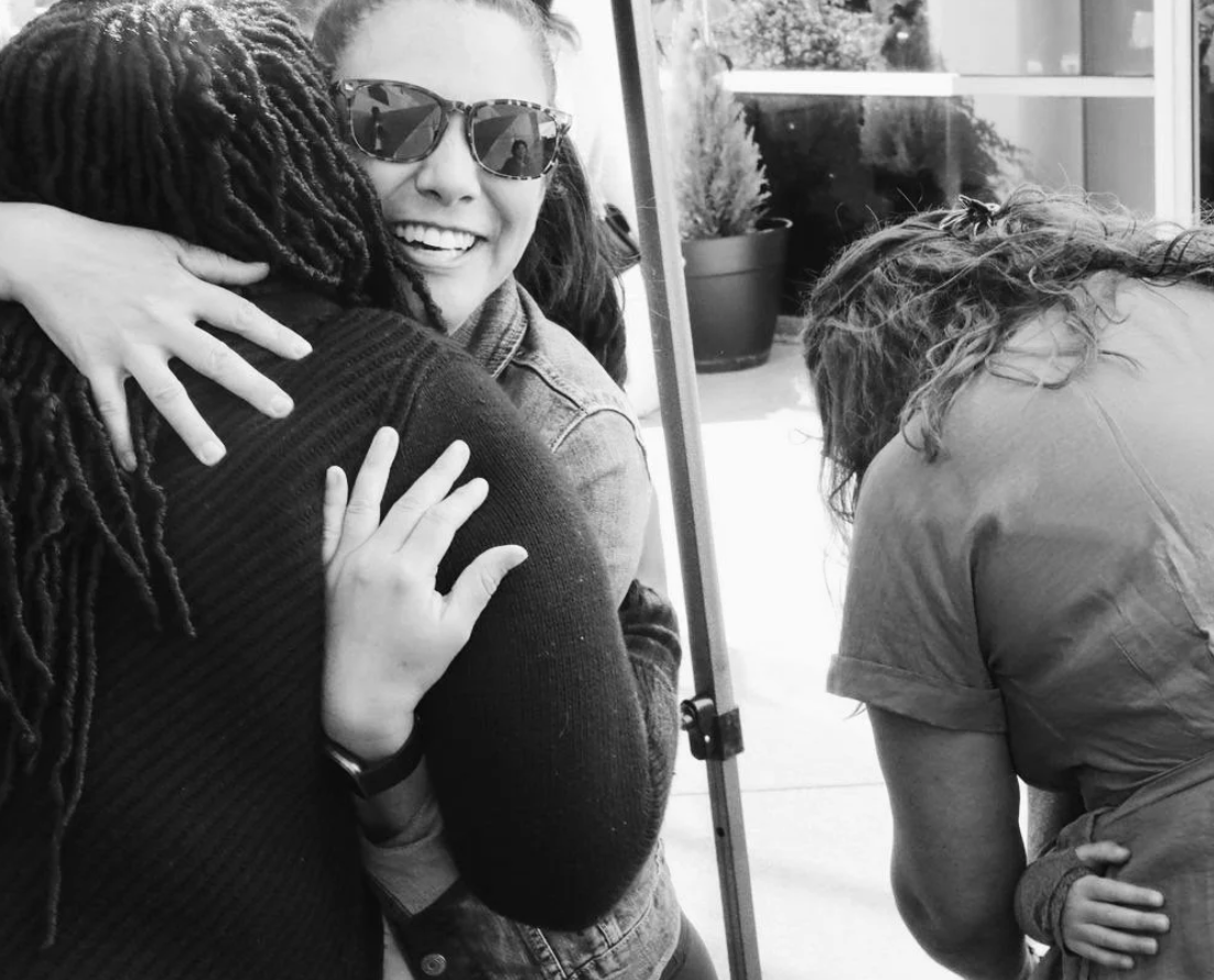 People hugging indoors, woman smiling wearing sunglasses, another person with curly hair, and a third person leaning over. Outdoor plants visible outside the window.