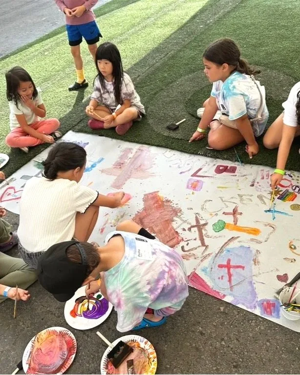 Kids drawing and painting on a large sheet of paper outdoors.