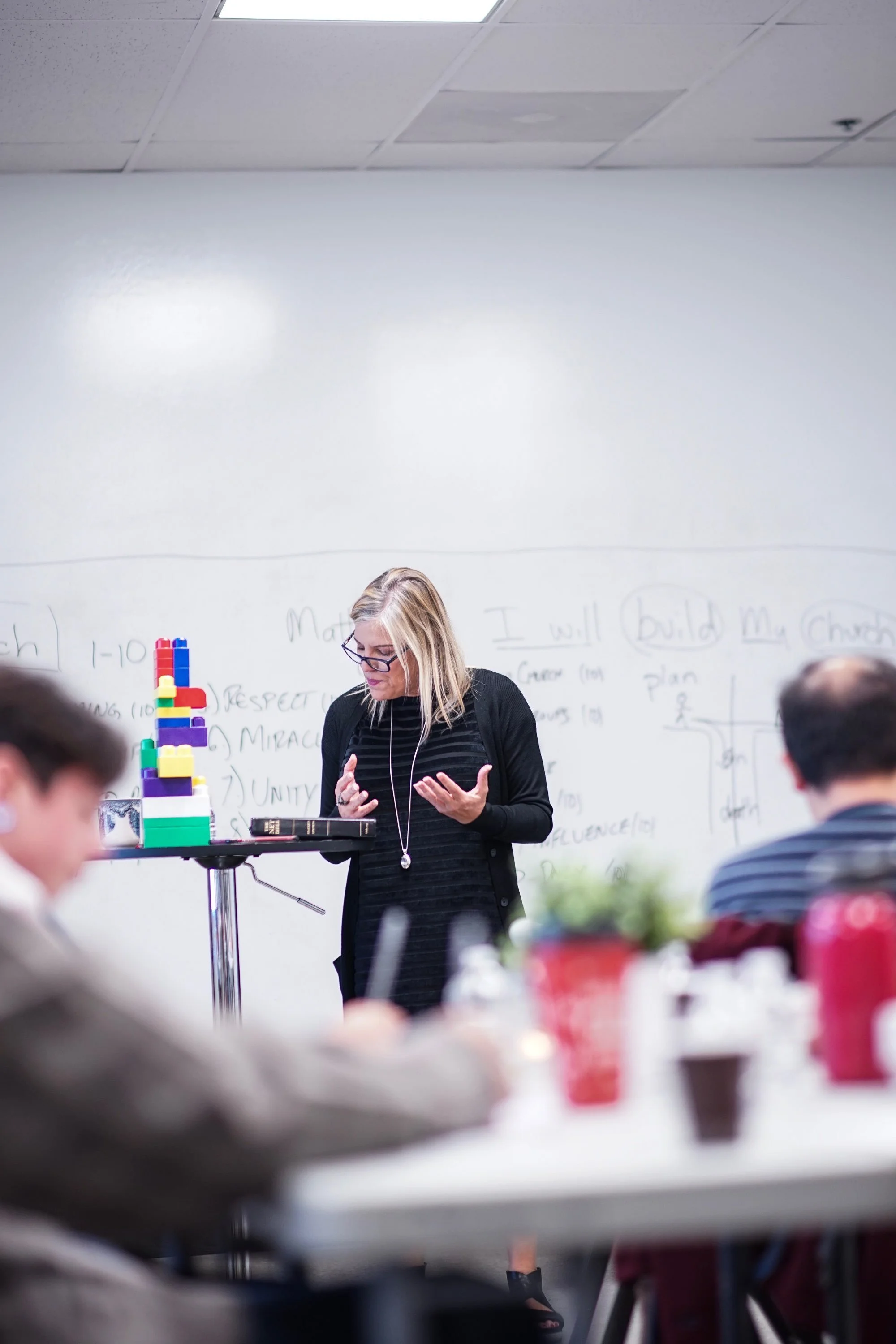 A woman in glasses and a black dress teaching in front of a classroom with students, a whiteboard, and a tower of colorful building blocks on a stand.