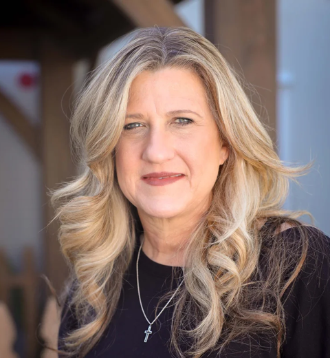 Simone Pascua. Children's pastor at Carmel Church in San Diego. She helps with the pre-school, kids church, and kids. Close-up of a middle-aged woman with long, wavy blonde hair, wearing a black top and a cross necklace, outdoors in natural lighting.
