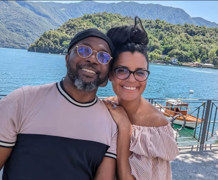 A smiling couple taking a selfie near a lake with mountains in the background. The man has sunglasses, a cap, and a beard. The woman has glasses and dark hair styled in a bun. They are standing close together, with the woman resting her hand on the man's shoulder. There is a docked boat and railing behind them.