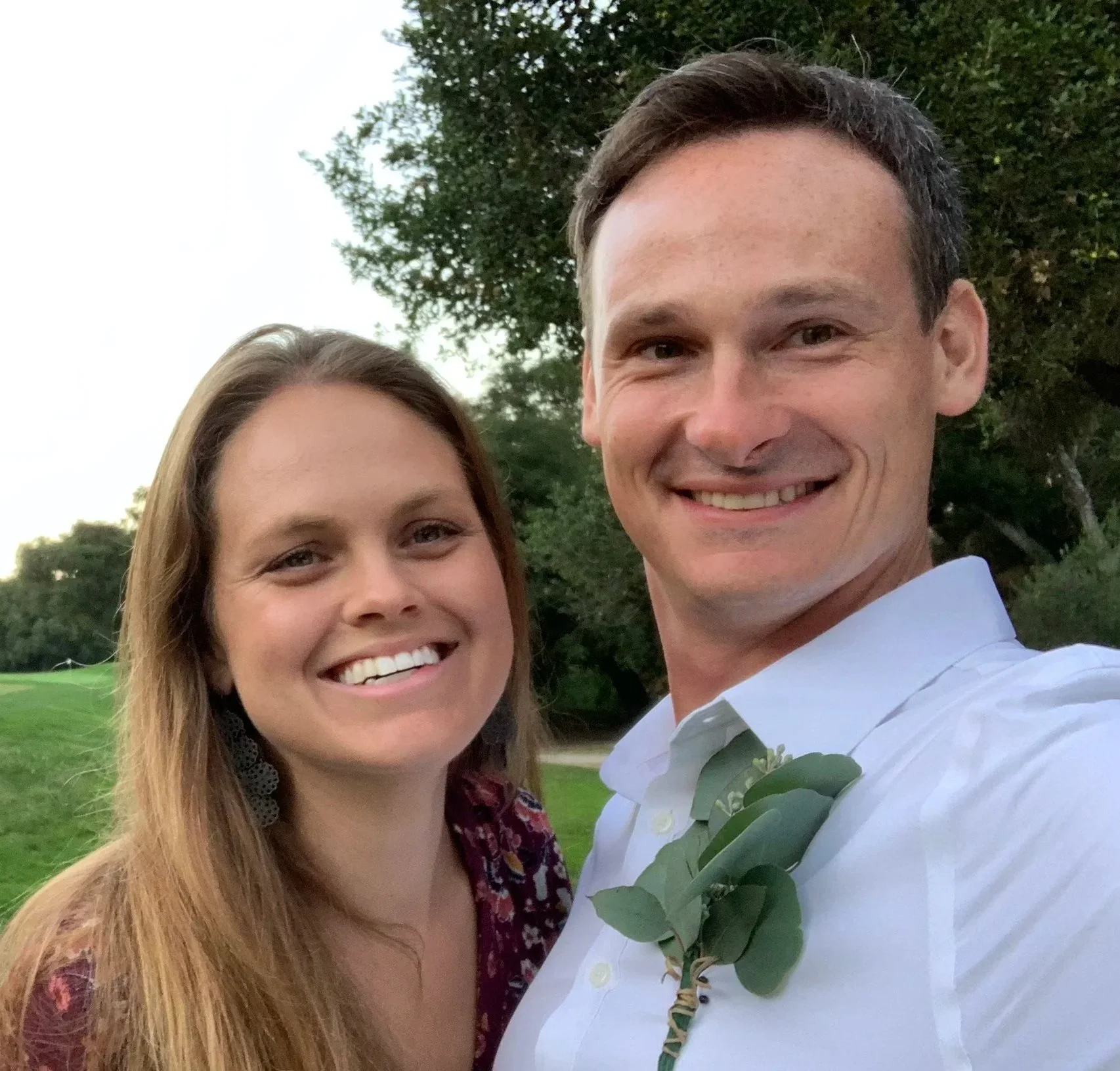 A smiling couple taking a selfie outdoors in a park with green trees and grass.