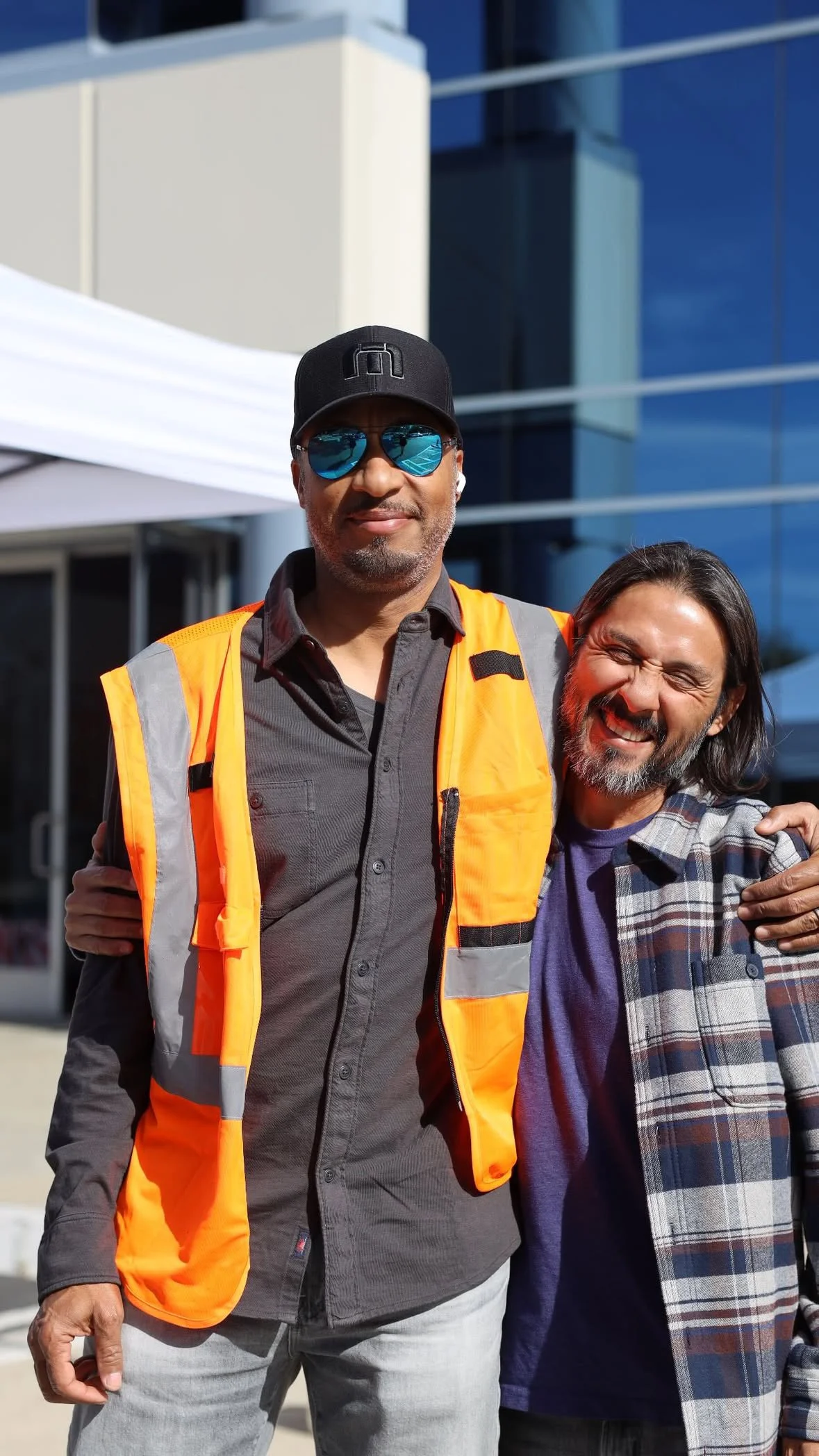 Two men smiling, one wearing a black cap, sunglasses, gray shirt, and an orange safety vest, the other wearing a plaid shirt and a jacket, standing outdoors in front of a modern building.