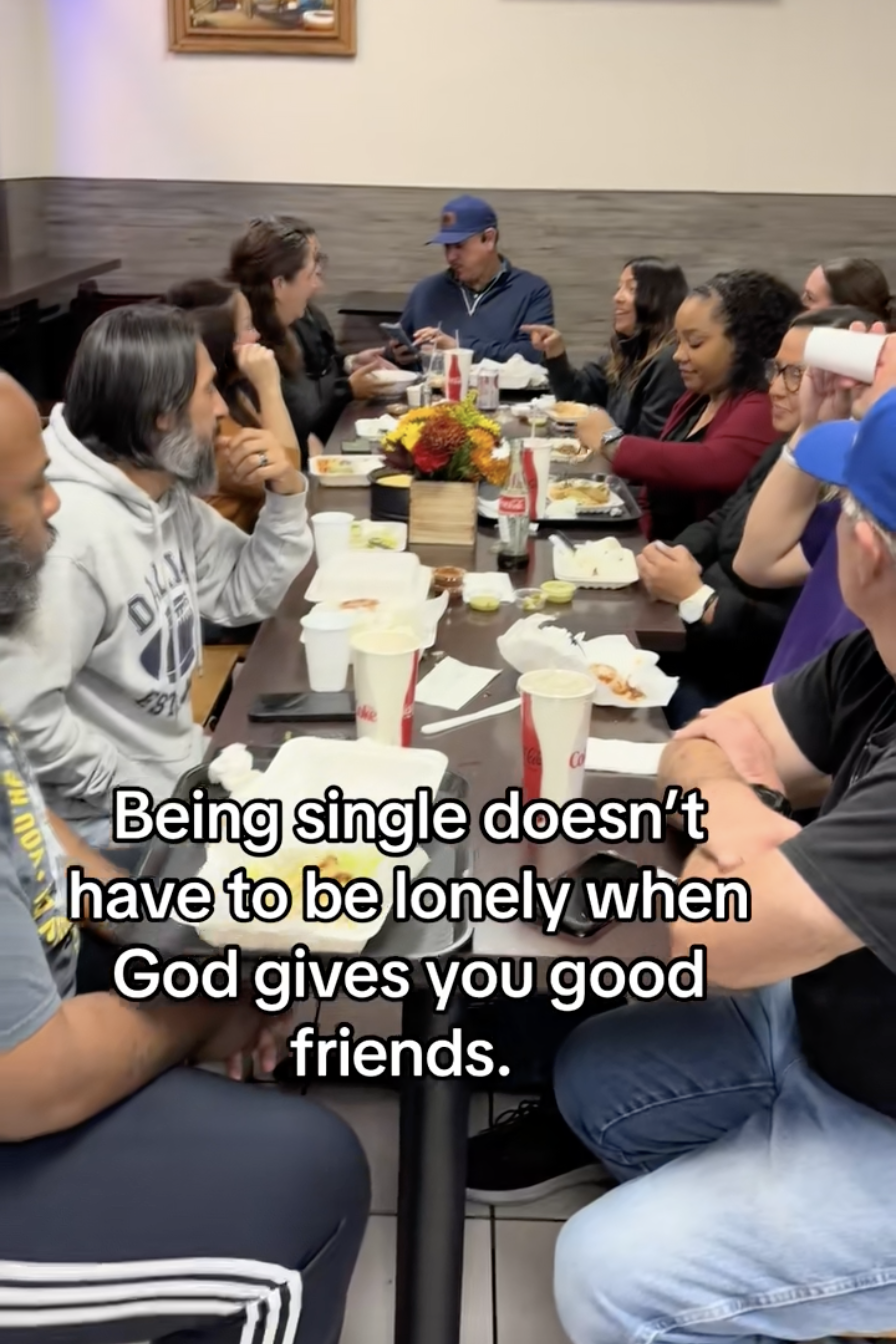A group of people sitting around a table, sharing a meal in a restaurant with some taken food containers and drinks on the table, and a flower centerpiece in the middle.
