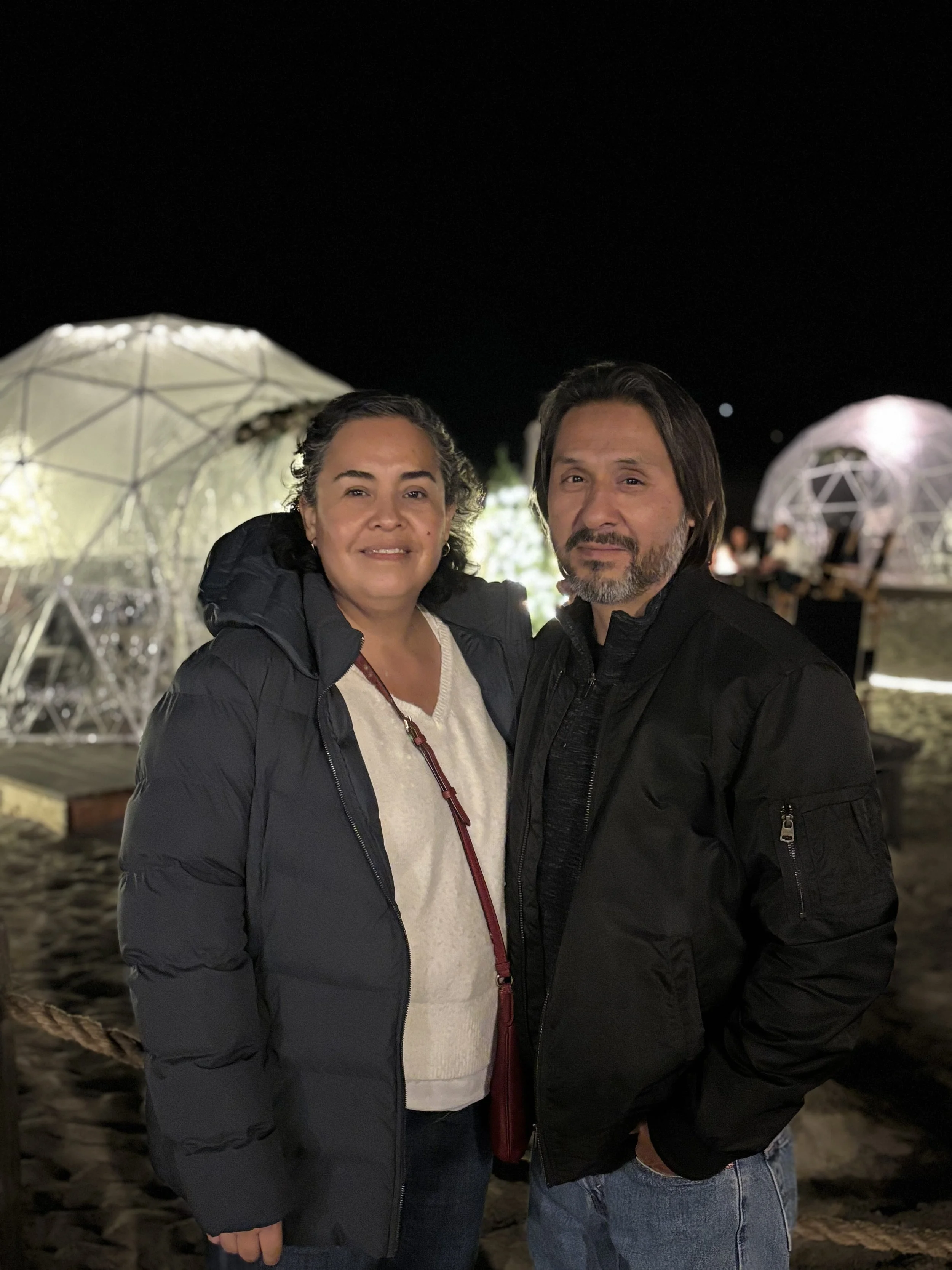 A woman and a man standing close together outdoors at night, with illuminated geodesic dome structures in the background.