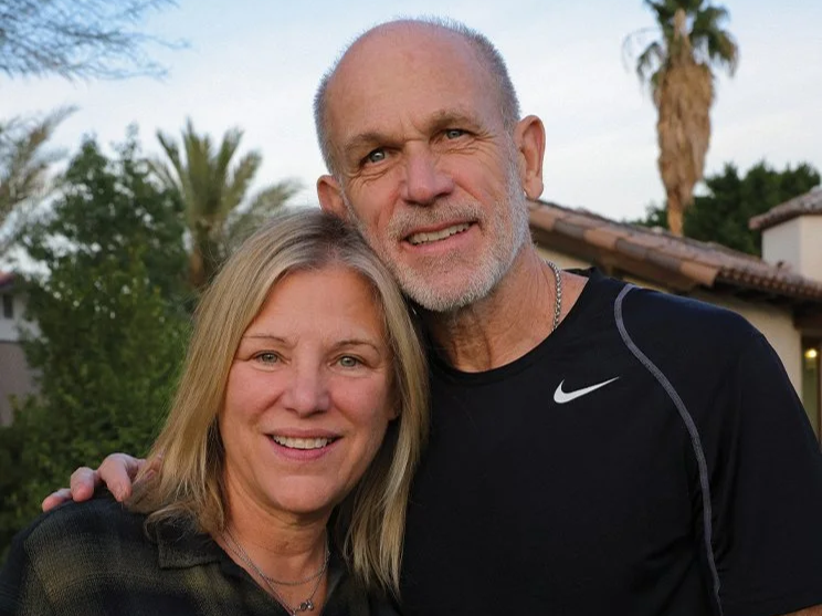 A middle-aged woman and an older man smiling together outdoors with trees and a house in the background.