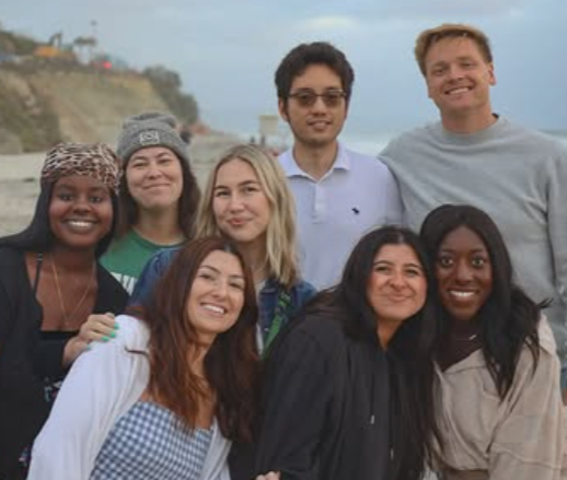 Group of seven young adults posing outdoors near a beach or coastal area.
