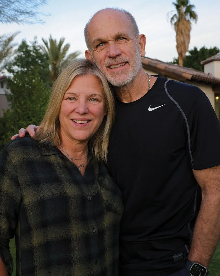 Smiling middle-aged woman with blonde hair and a middle-aged man with a beard, both wearing casual clothing, standing outdoors with palm trees and houses in the background.