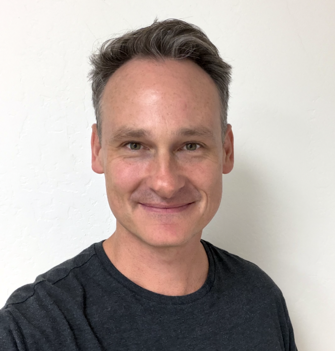 Nic Schneider is the college pastor at Carmel Church in San Diego. He is on Palomar college, san diego state, cal state san marcos, etc. A smiling man with short, dark hair and light skin, wearing a black t-shirt, standing against a plain white wall.
