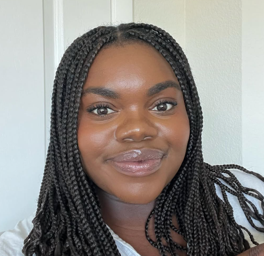 Menza Bolamba is the 4th-5th grade pastor at Carmel Church in San Diego. Close-up of a woman with dark braided hair, smiling softly, with a light-colored wall in the background.