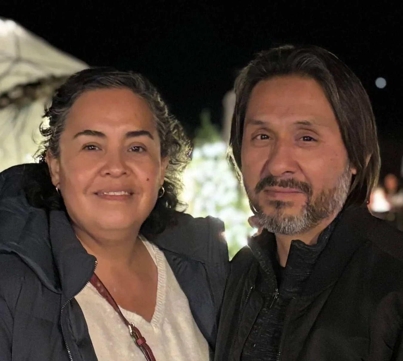 Mario and Rocio Robles are the Single Adults pastors at Carmel Church in San Diego. They help single parents build community in Jesus. A man and woman posing together at night, smiling, with a blurred background of outdoor lights and possibly tents.