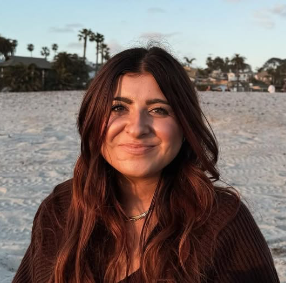 Maddy Rodriguez is on staff at Carmel Church in San Diego. She helps in creative arts, worship, Instagram, social media, and more. A woman with long wavy brown hair smiling at the beach, with palm trees and houses in the background during sunset.