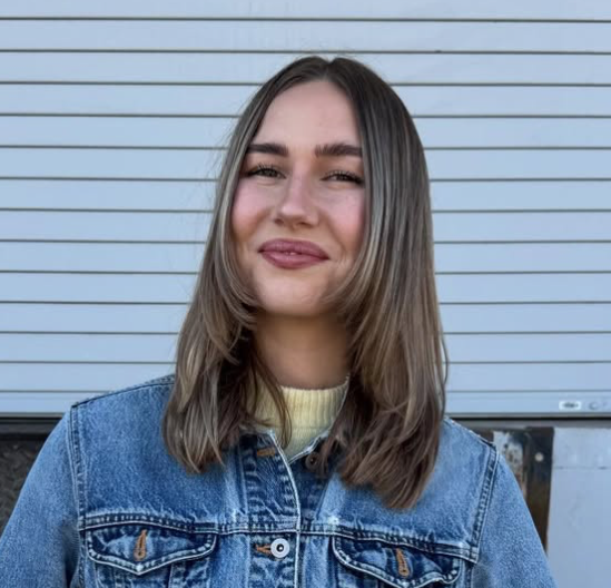 Kassidy Hard is the High School pastor at Carmel Church in San Diego. A young woman with shoulder-length brown hair, smiling with closed eyes, wearing a denim jacket and standing in front of a light gray, horizontally-slatted wall.