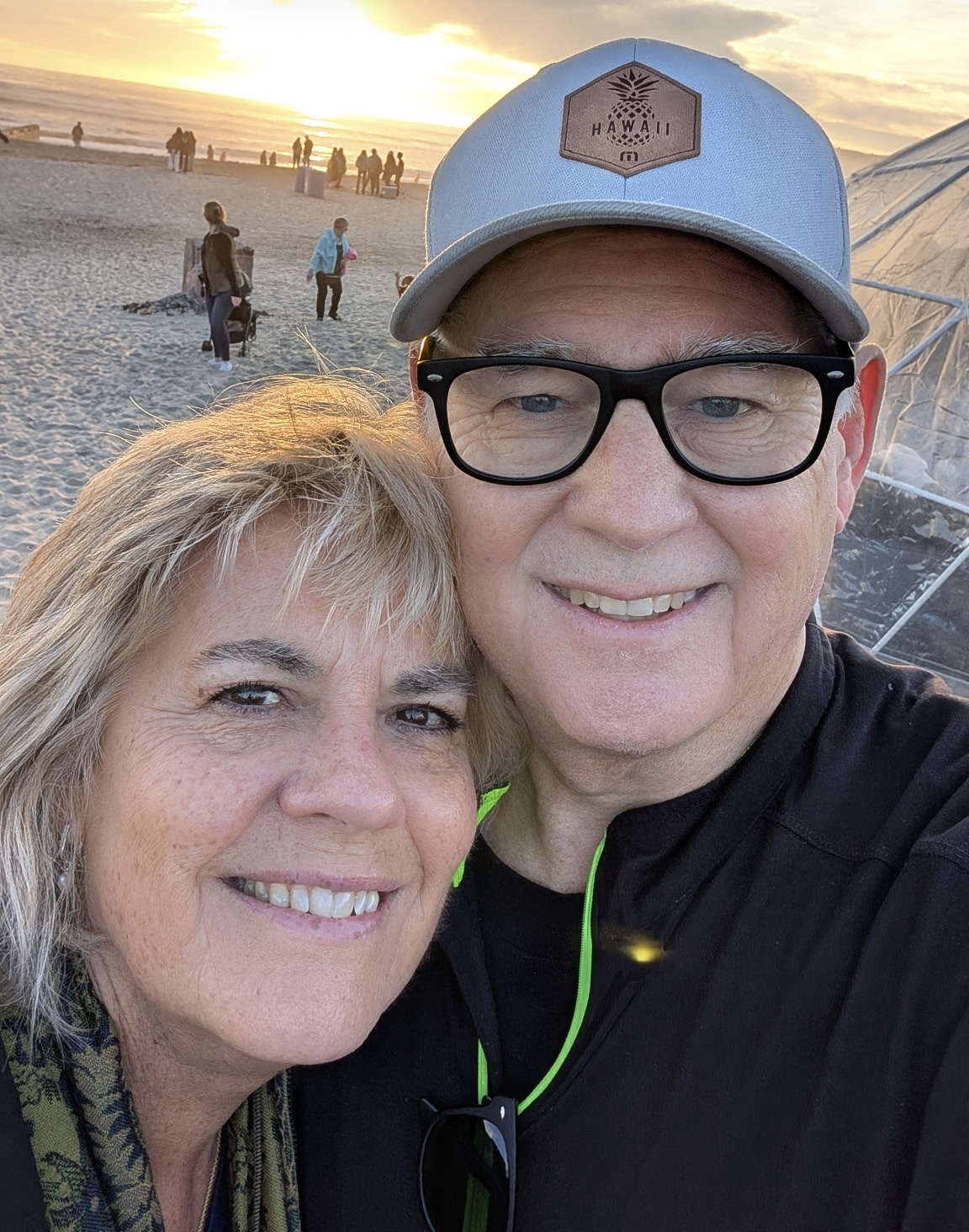 Dan and Kim Roy are the Adults pastors at Carmel Church in San Diego. They help build community who loves Jesus. A smiling couple taking a selfie at the beach during sunset, with the ocean and other people in the background.