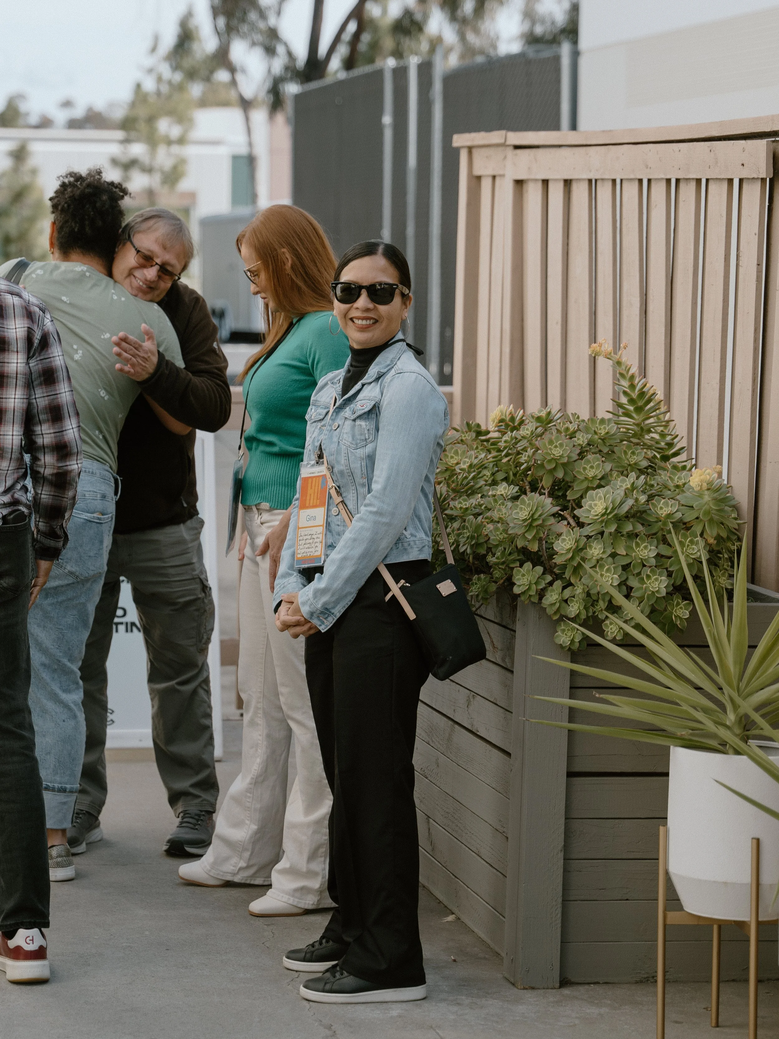 Carmel Church is a friendly church. Make friends, love Jesus, build community, walk close to God, build confidence, experience miracles. A group of people standing outdoors, smiling and interacting near a wooden fence and plants.