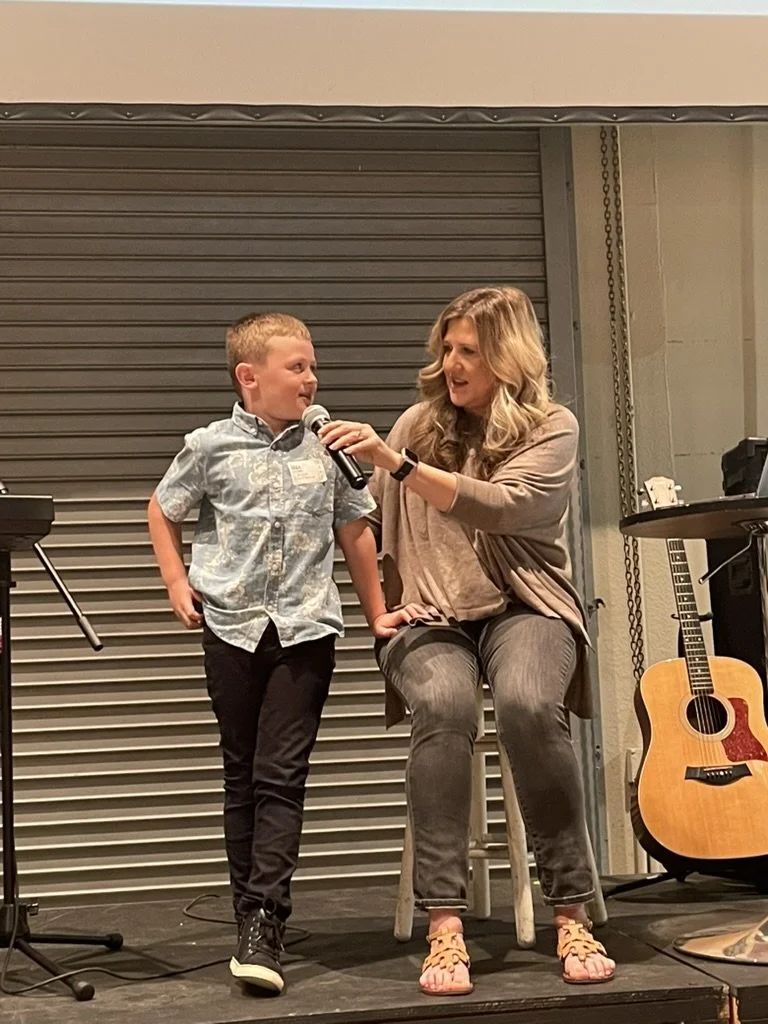 A young boy is speaking into a microphone held by a woman sitting next to him on a stage, with a guitar and keyboard nearby.