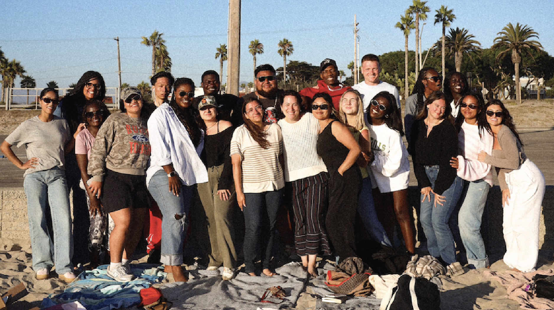 A large group of diverse people posing together on the beach with palm trees and power lines in the background, smiling and enjoying the sunny day.