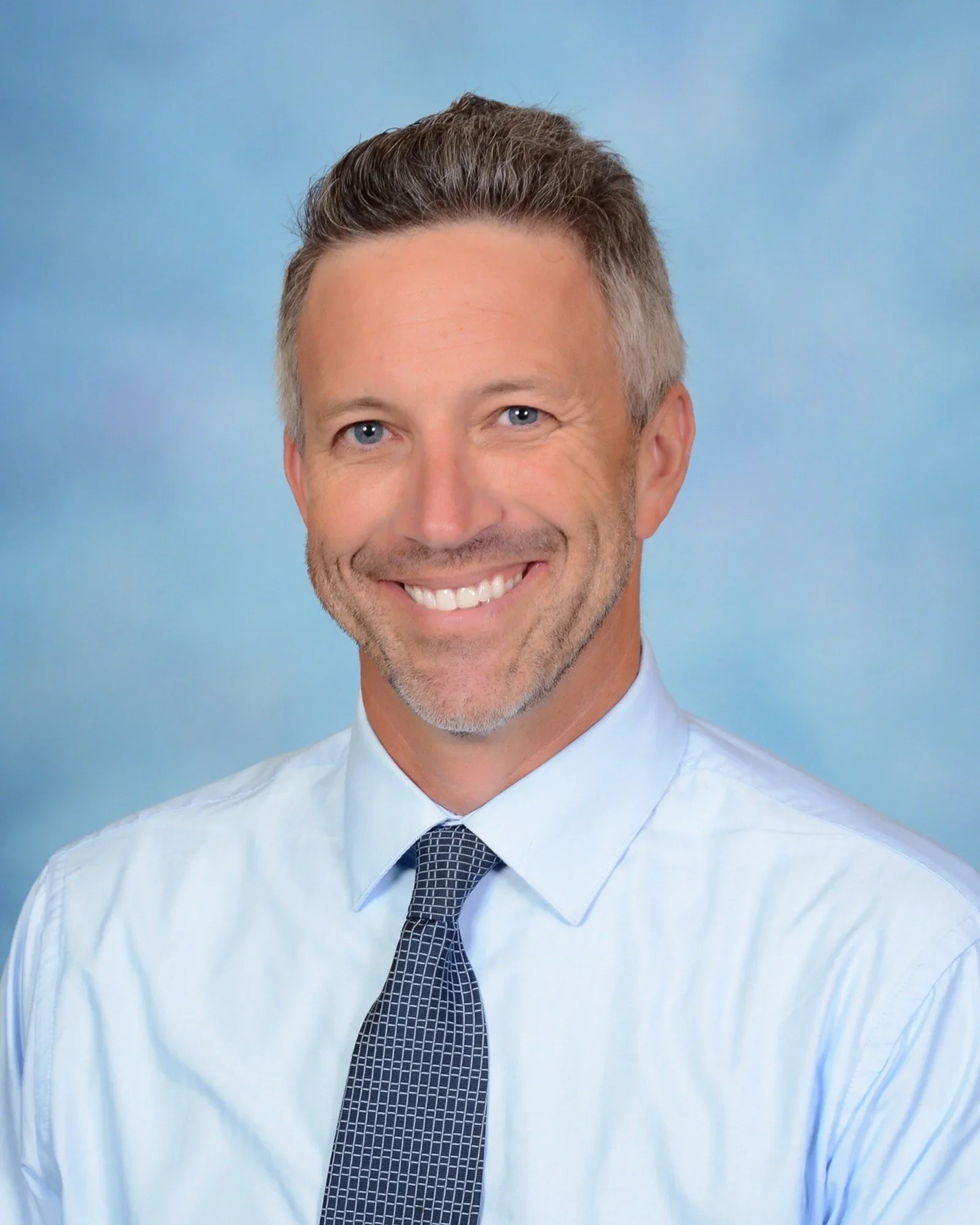 Headshot of a smiling man with styled hair, light stubble, wearing a light blue shirt and a patterned blue tie, against a dark blue background.