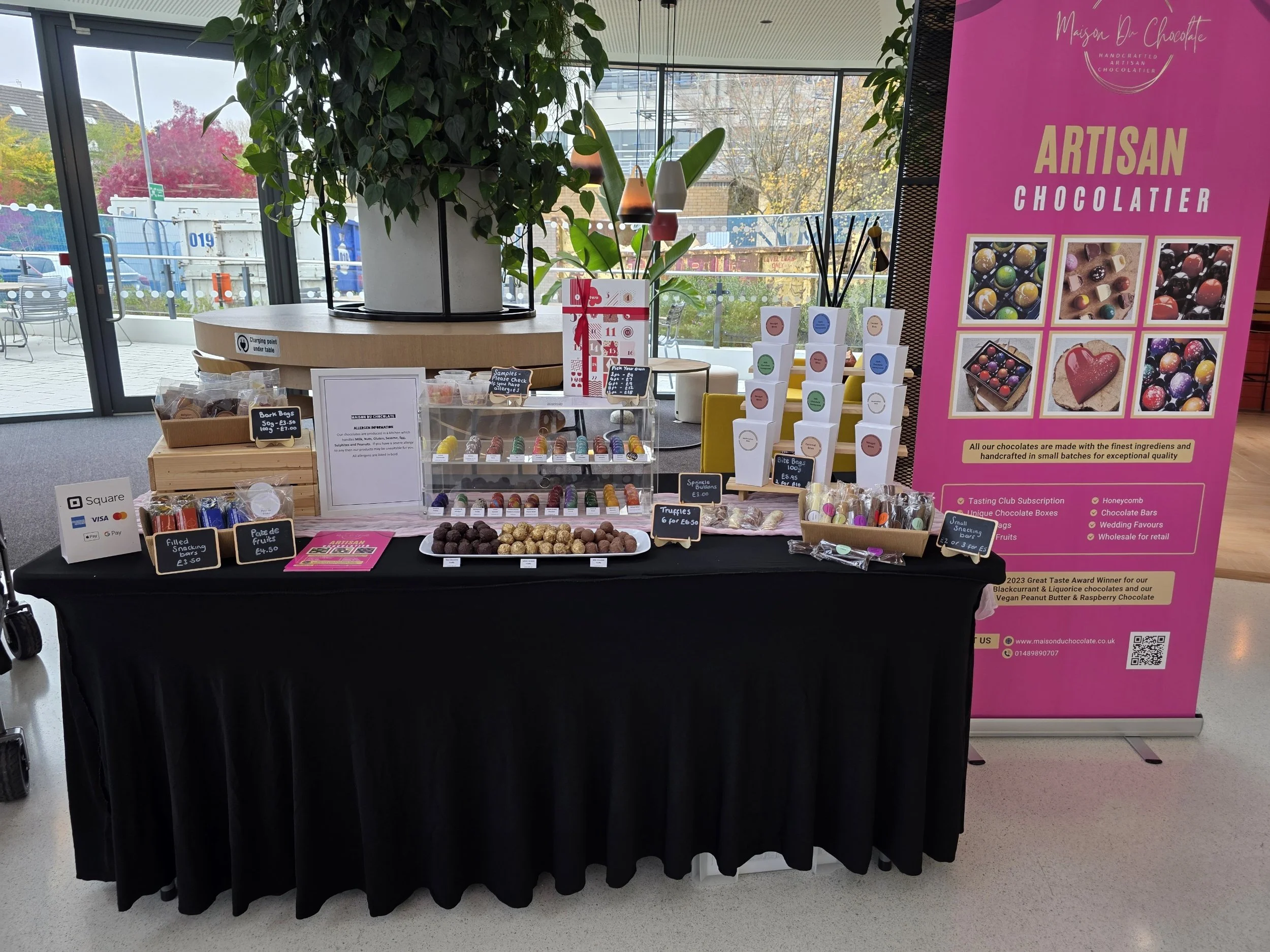 Display table with colorful chocolates, truffles, and candies for sale, branded with a pink banner that reads 'Artisan Chocolatier.' The setup includes small signs with prices and labels, with a large potted plant on the left and large windows in the background.