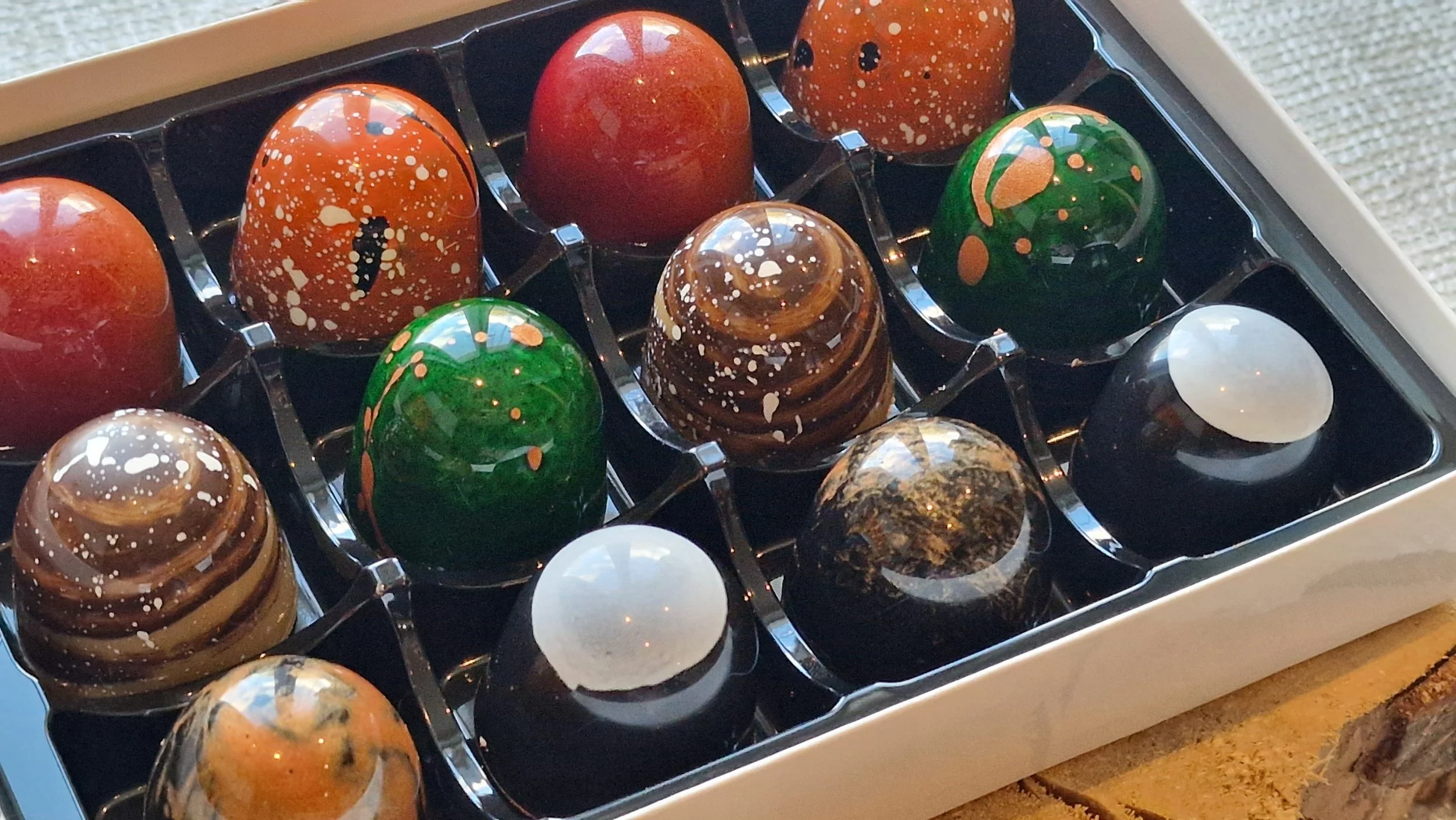 A box of assorted colorful chocolates with various marbled and splattered designs, arranged in a black plastic tray.