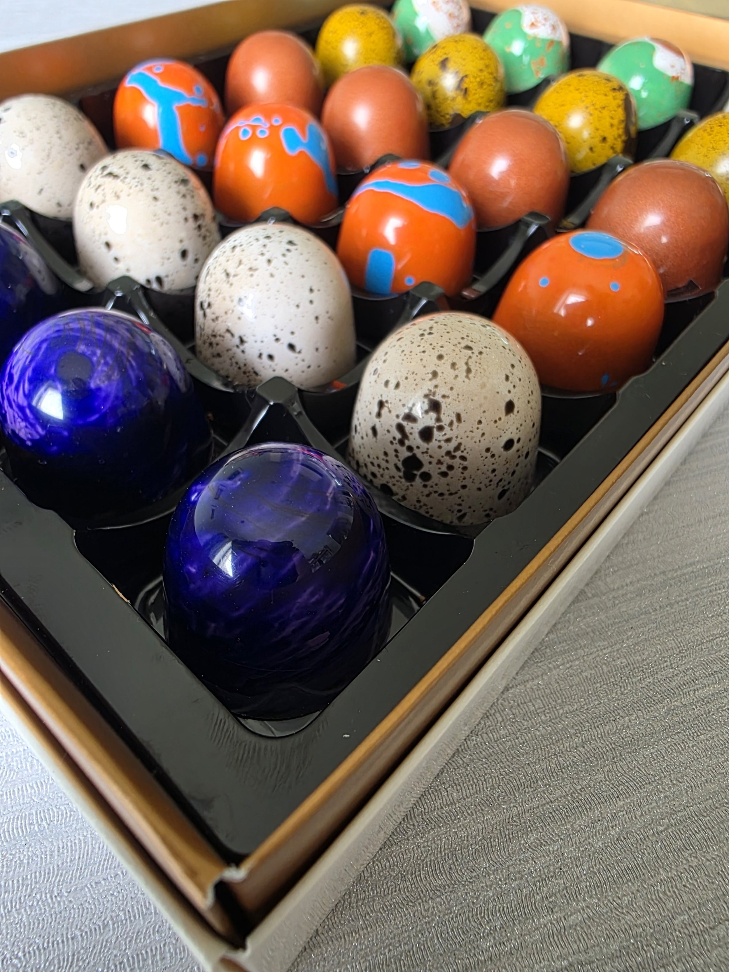 Decorative eggs in a box, including speckled, orange with blue designs, green with white and pink, and navy blue marble patterns.