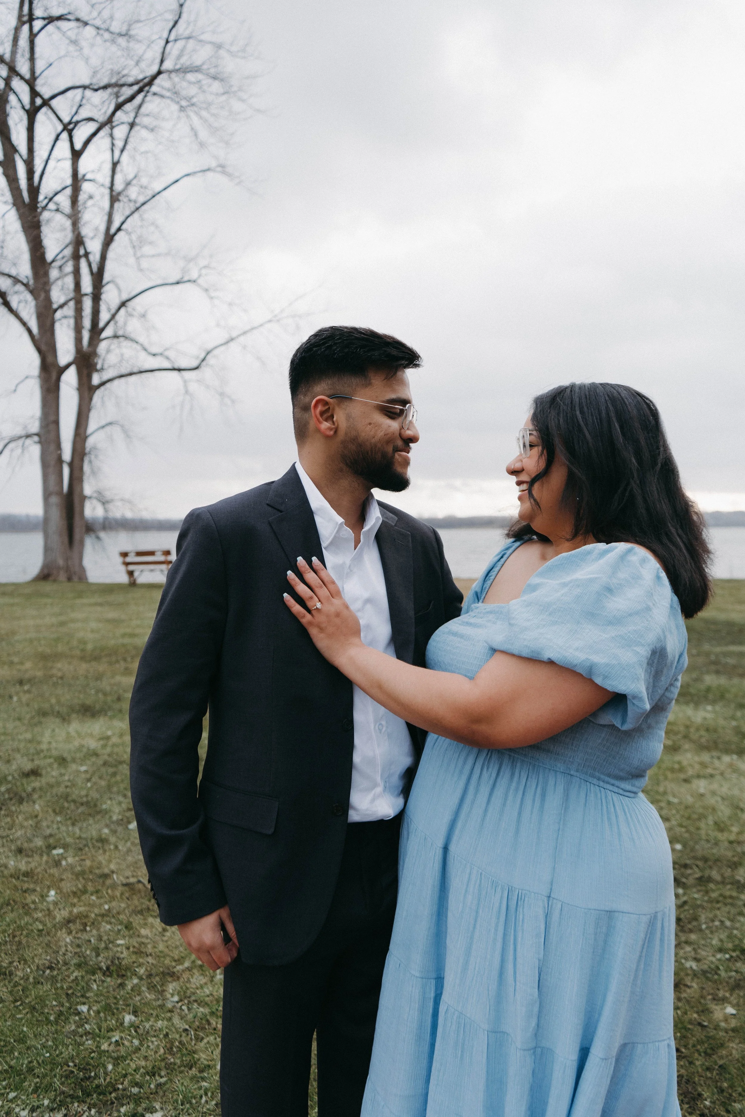 A couple standing close together outdoors near a lake, with trees in the background. The man is wearing a dark suit and white shirt, and the woman is in a light blue dress. They are smiling and looking at each other affectionately.