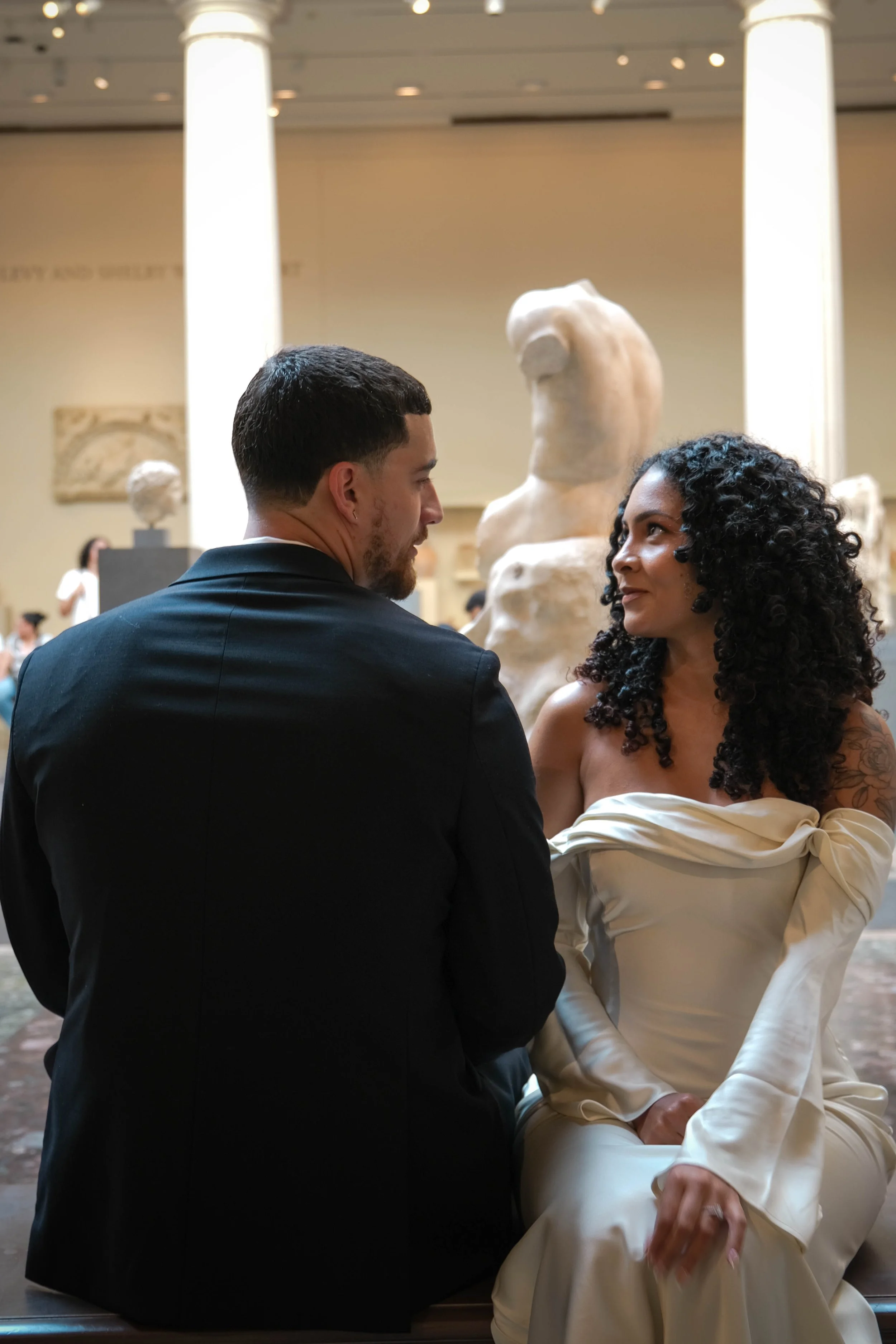 The Met Engagement Shoot A man and woman sitting close together and looking at each other in an art museum, with classical sculptures in the background.