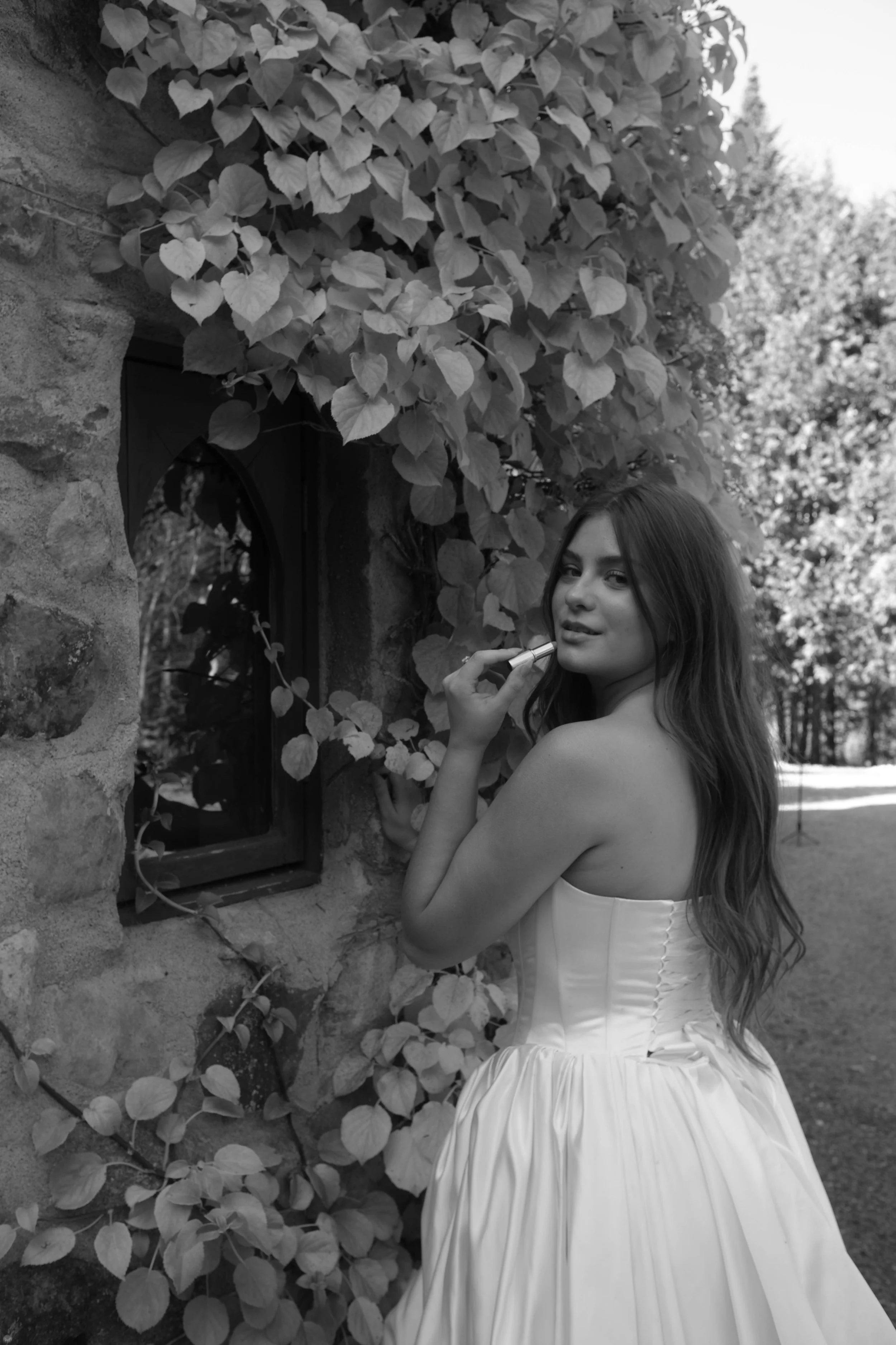 Irasburg, Vermont A woman in a strapless white dress standing outdoors near a wall with ivy, holding a lipstick near her face, with trees and sunlight in the background.