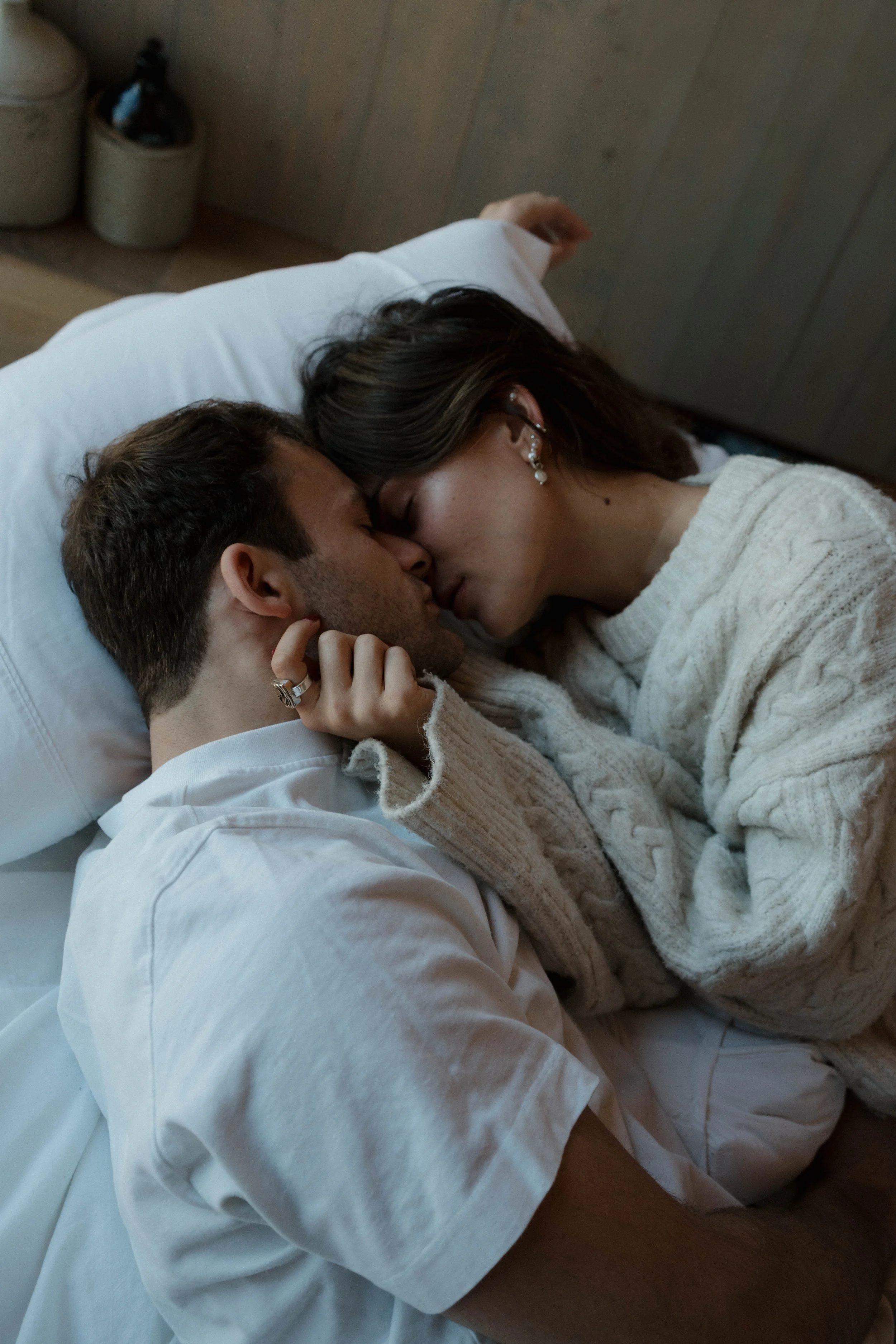 A couple lying in bed, sharing a kiss, with the woman gently holding the man's face and their noses touching.